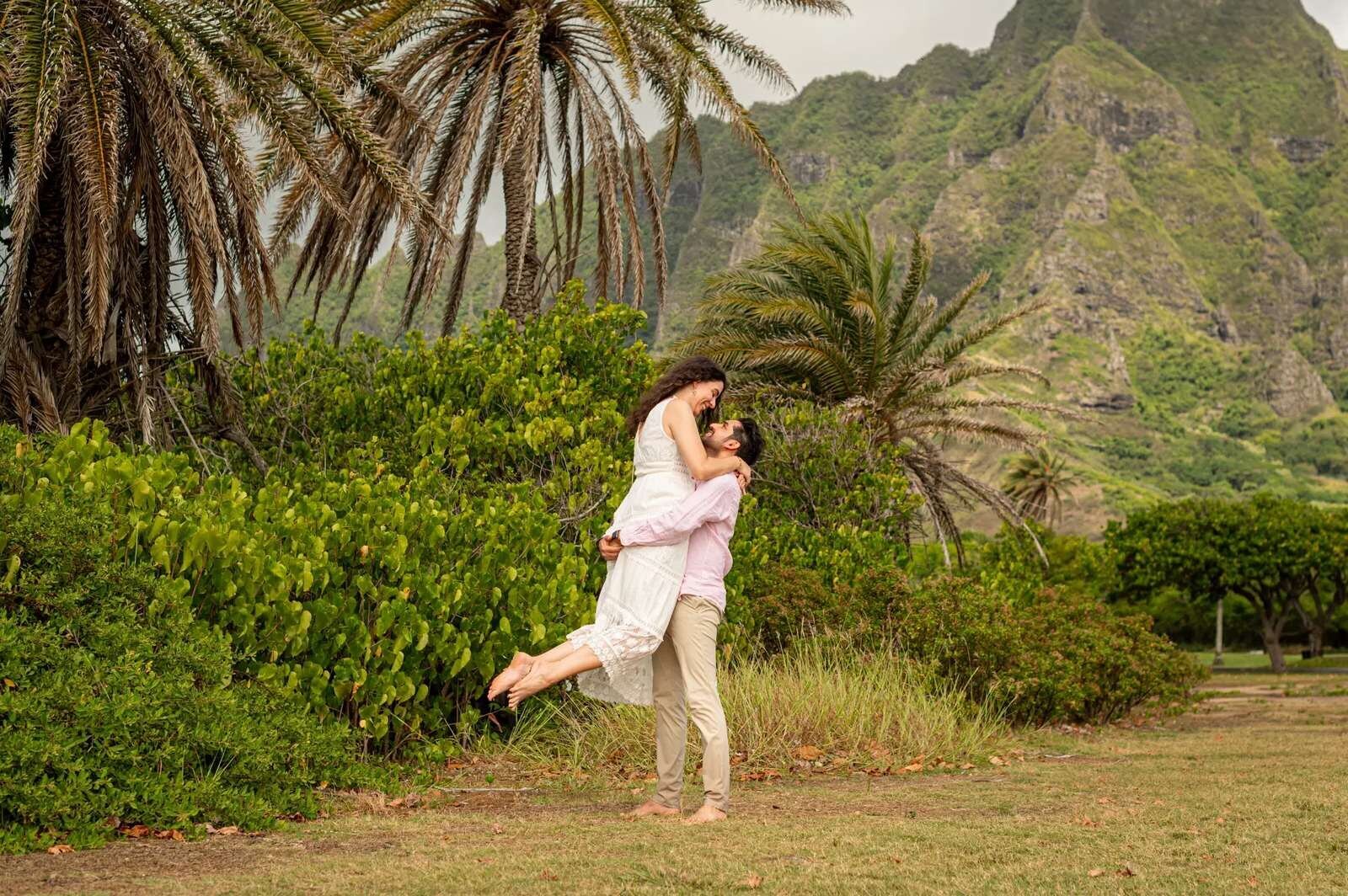 kualoa-engagement-photos-oahu-desktop A couple on a grassy lawn, the man lifting the woman in a white lace dress, with palm trees and jagged green mountains behind them
