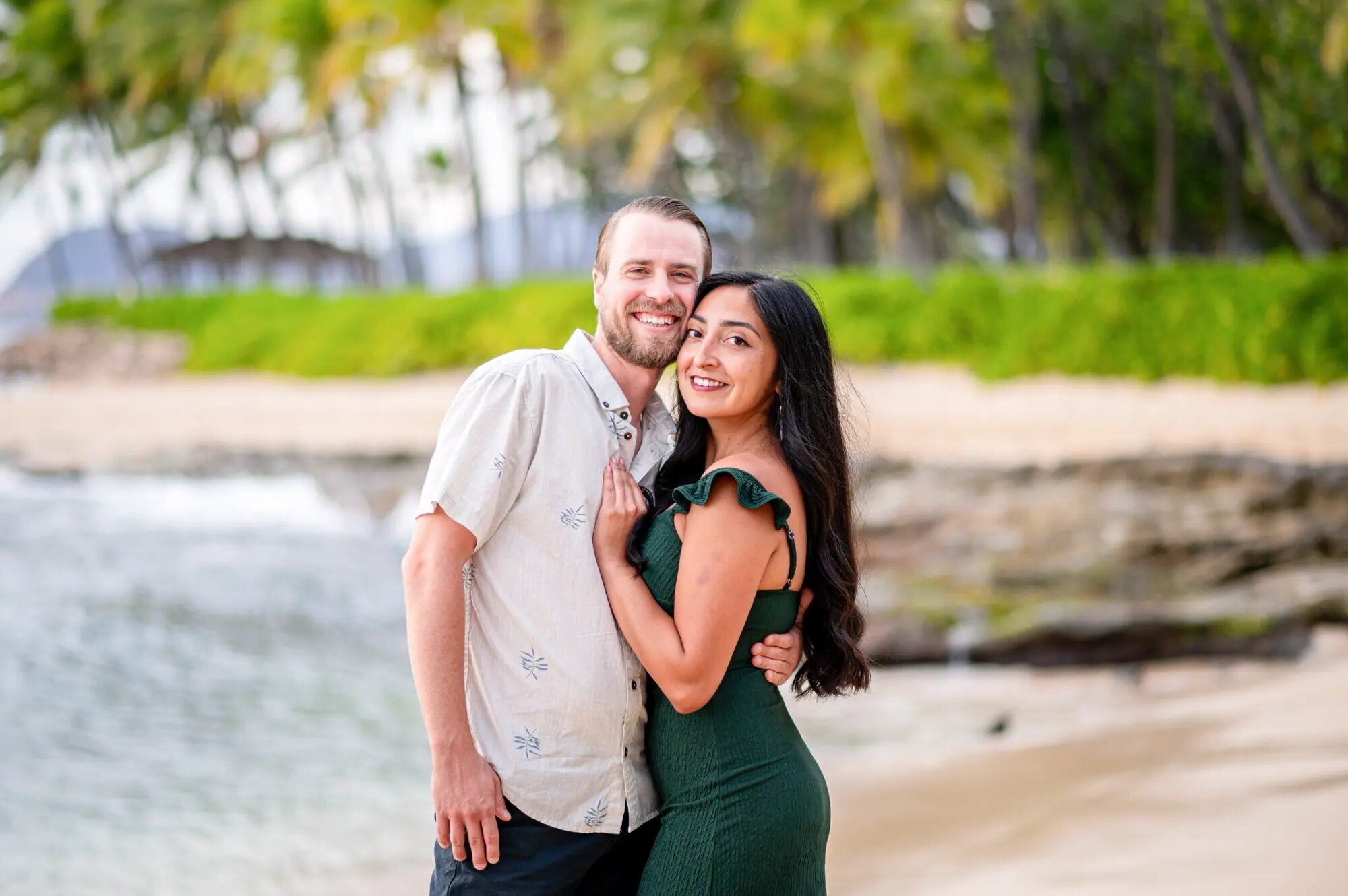Smiling couple embracing on Ko Olina Beach with tropical foliage and calm water behind them