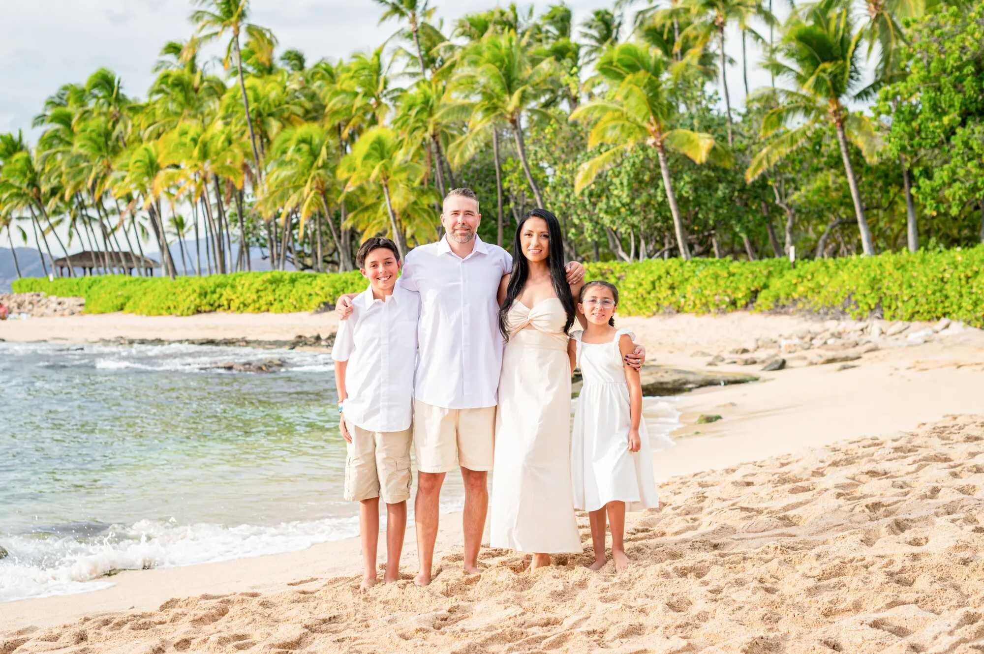 A family of four in white and cream outfits posing together on the sandy shore at Ko Olina Beach on Oahu with lush green palm trees and calm turquoise water behind them