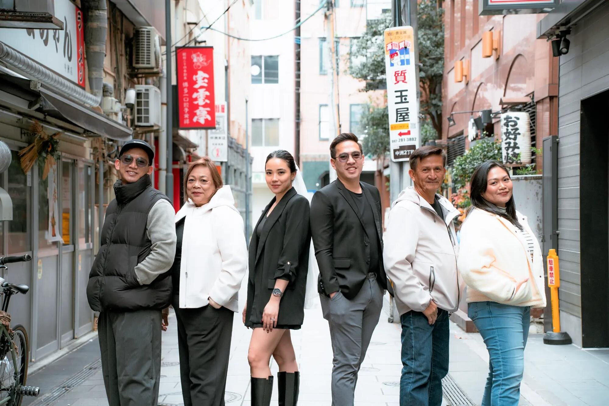 Fukuoka Family Photography — Nakasu Street Session | Dreamgraphy A stylish multigenerational family posing together on a narrow Nakasu alley lined with Japanese shop signs in Fukuoka. A relaxed, editorial-style family photo session captured by Dreamgraphy.