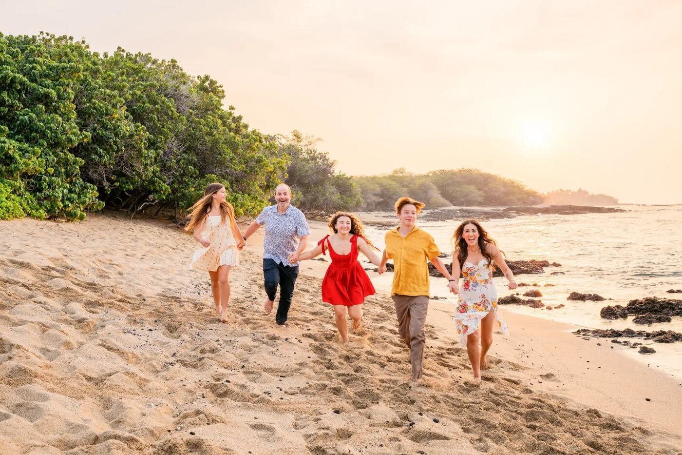 Big Island Family Photos — Candid Beach Sunset Session Kona A family of five laughing and running together along a Big Island beach at sunset with lush trees and ocean in the background