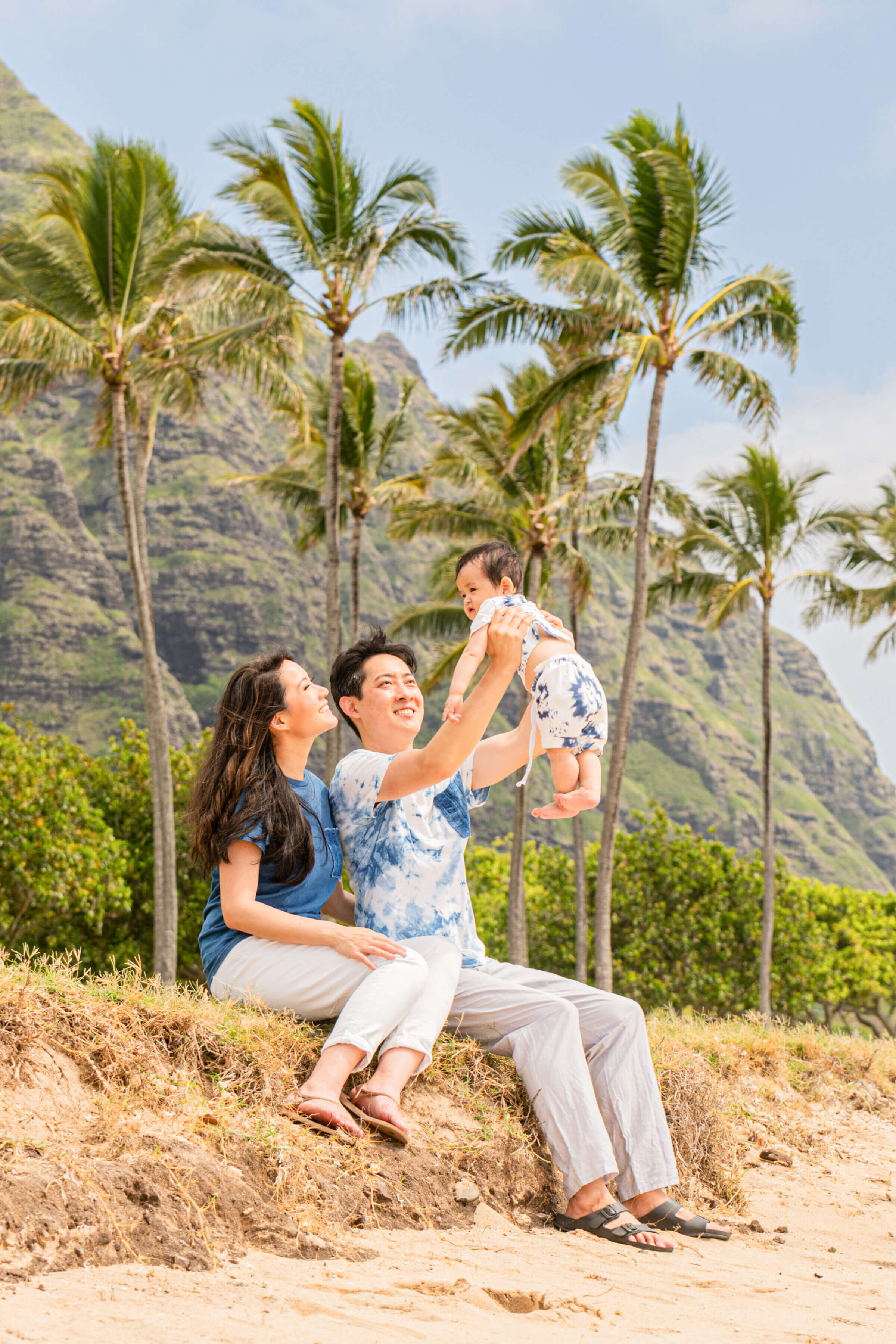 Hawaii Family Vacation Photographer — Parents with Baby Against Dramatic Mountain Cliffs A couple sitting on a sandy hillside lifting their baby in the air and laughing during a family vacation photography session in Hawaii, with tall palm trees and dramatic green mountain cliffs in the background