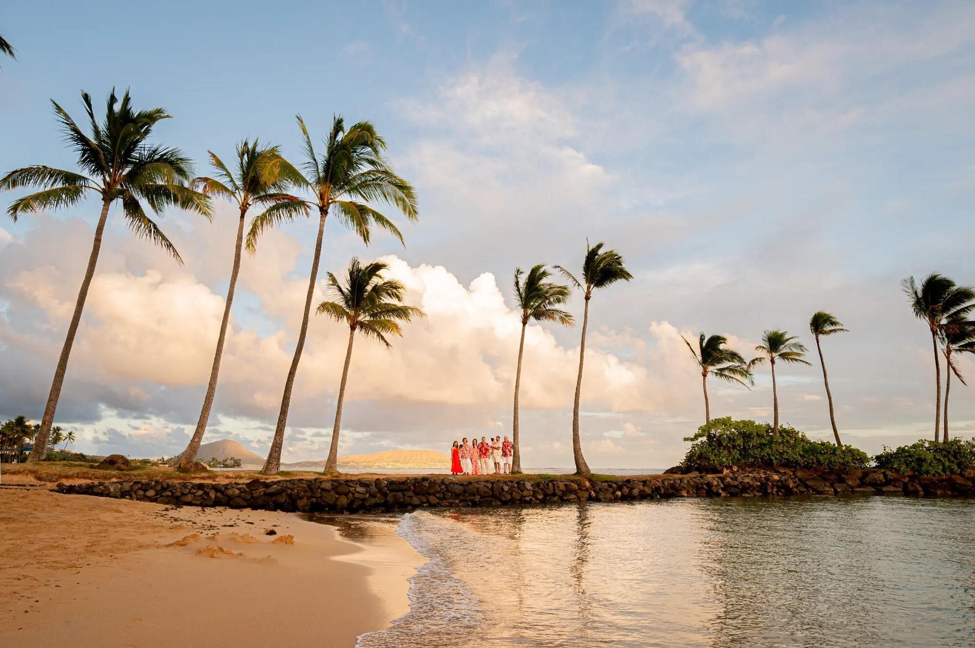A family group posing on a stone jetty at Waialae Beach on Oahu surrounded by tall swaying palm trees with golden sunset light and calm ocean water reflecting the sky