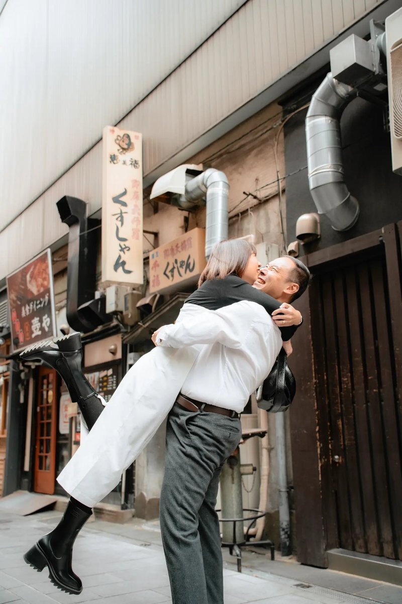 fukuoka-photographer-location-dazaifu-mobile A laughing man lifts a woman in black boots off the ground in a narrow Fukuoka street lined with Japanese restaurant signs and metal ducts