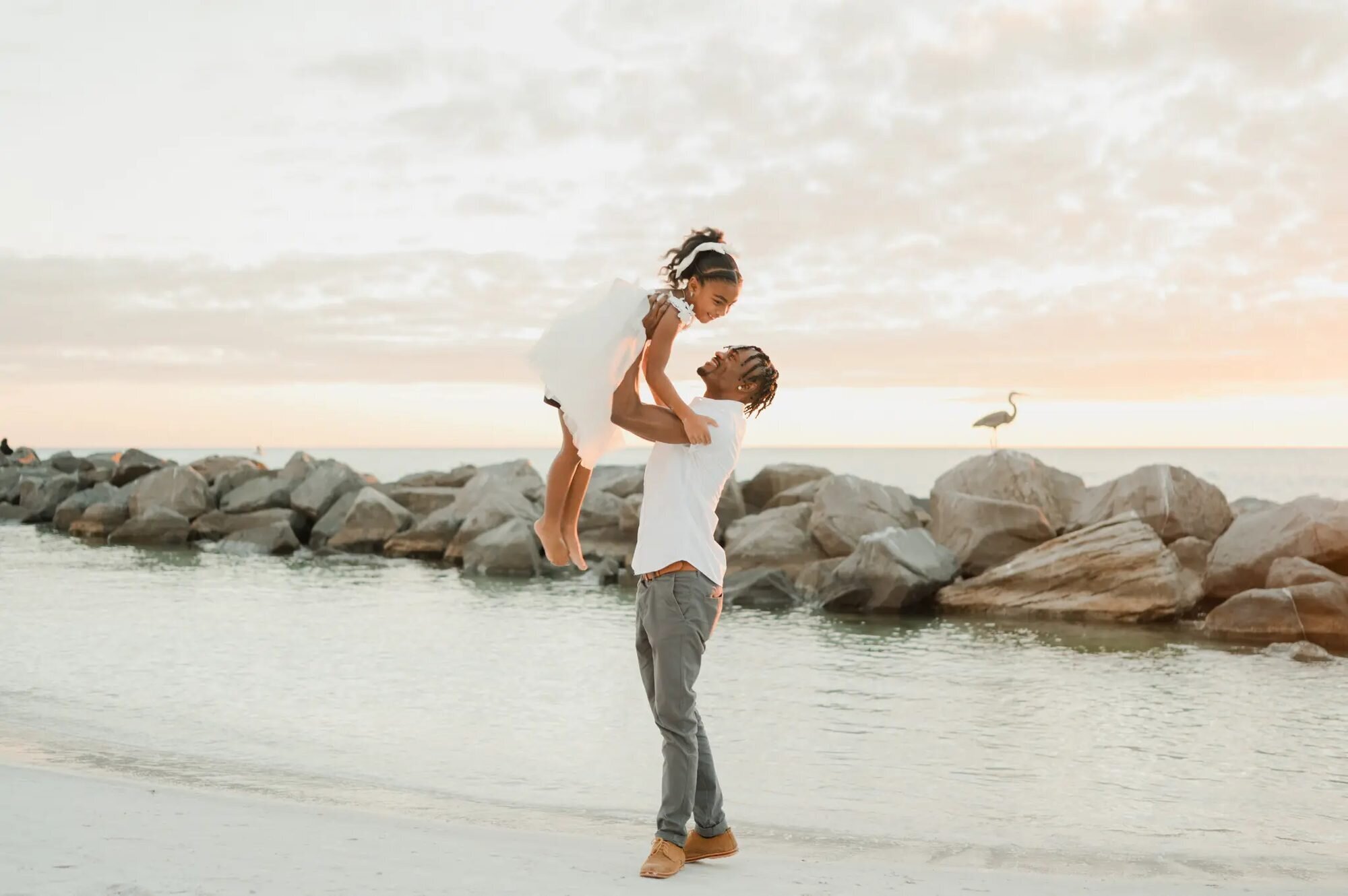 Tampa Family Photographer | Father & Daughter Session at St. Pete Beach A father lifting his young daughter joyfully in the air on the shoreline at St. Pete Beach, Tampa, with rocky jetties and a great blue heron in the background — captured by a Dreamgraphy Tampa family photographer