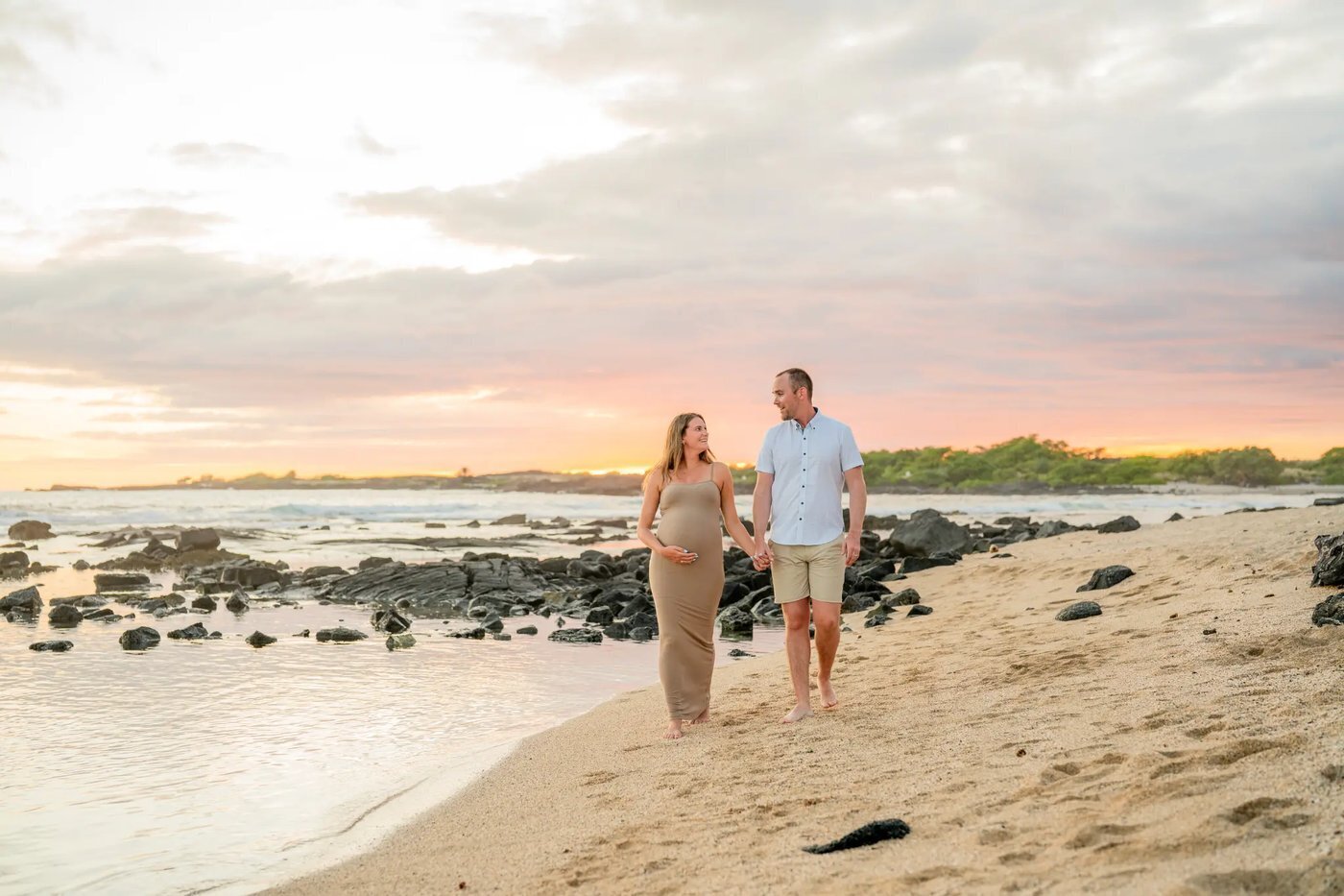 Maternity Photos Big Island Hawaii — Beach Sunset Session Kohala Coast A pregnant woman in a tan dress holding her belly while walking hand in hand with her partner on a Big Island beach at sunset with lava rocks and ocean in the background