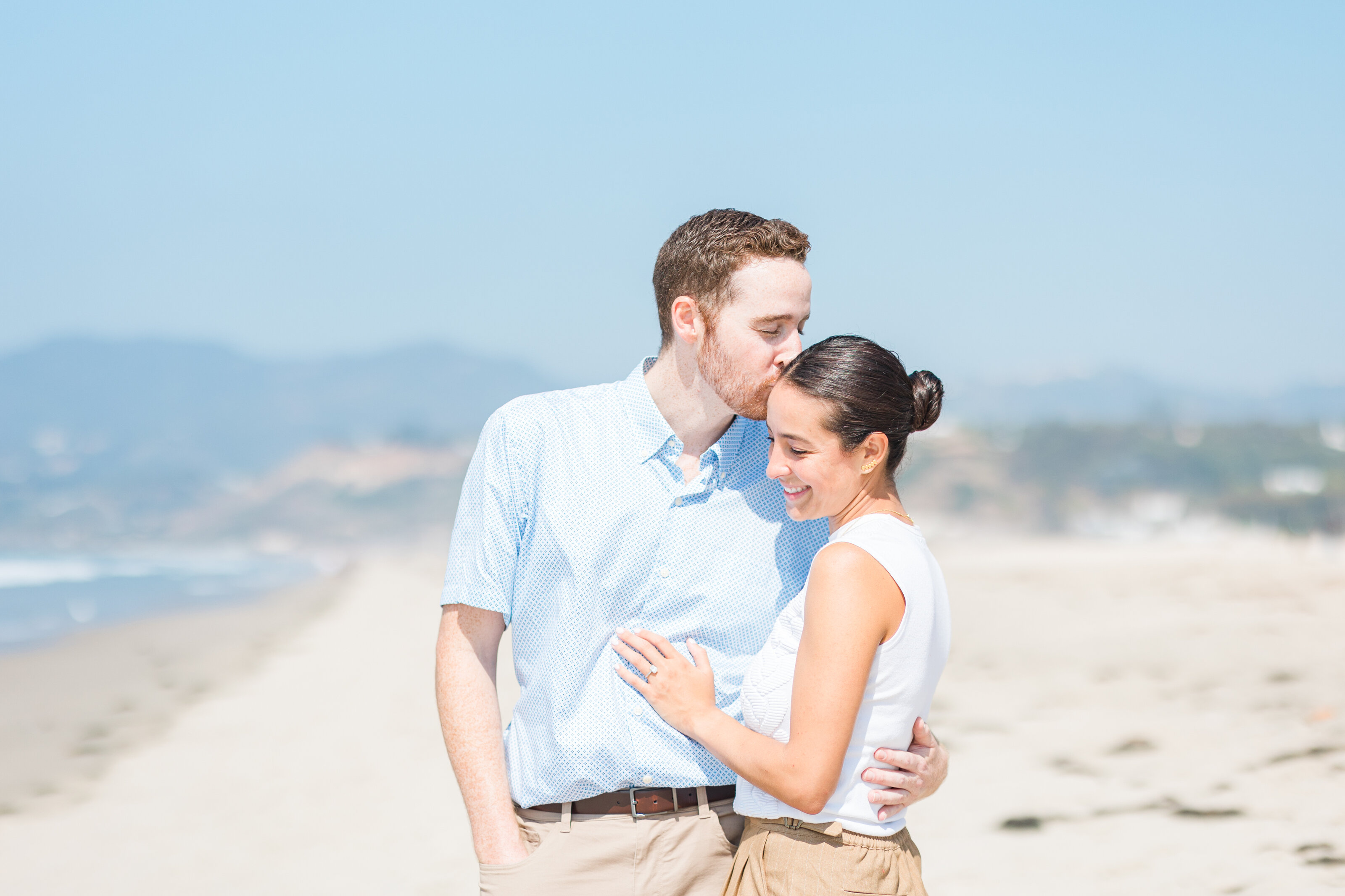 los-angeles-photographer-couples-session-santa-monica-beach-mobile A man kissing the forehead of a smiling woman as they embrace on a sunny beach in Los Angeles, with hazy hills and the ocean behind