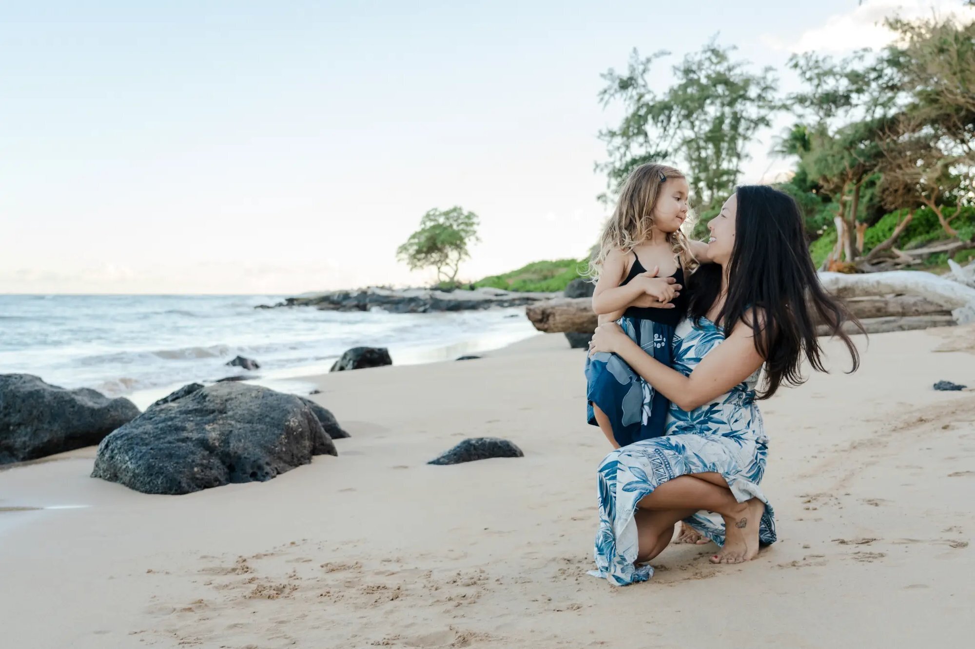 Kauai Photographer — Family Beach Photo Session | Dreamgraphy A mother crouching on a sandy Kauai beach holding her young daughter and smiling during a professional family photo session, with ocean waves and tropical trees in the background