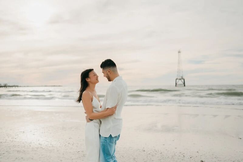 Tampa Photographer | Couple Session at Clearwater Beach A couple embracing and laughing on the white sand beach at Clearwater Beach, Tampa, with a pier and calm Gulf waters in the background — captured by a Dreamgraphy Tampa photographer