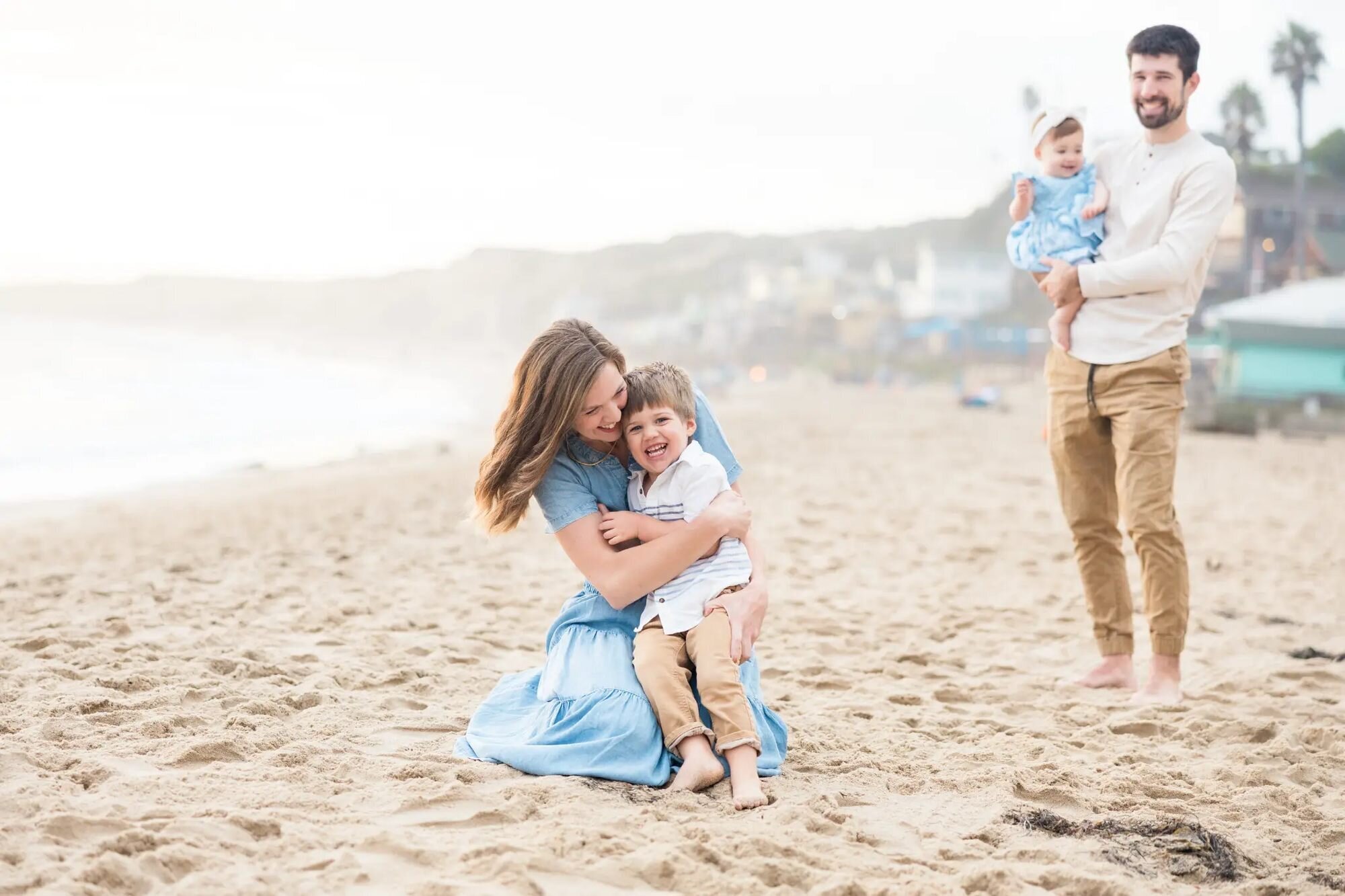 Los Angeles Photographer — Family Session at Seal Beach Los Angeles photographer capturing a mother hugging her young son on the sand at Seal Beach while the father holds their baby daughter in the background