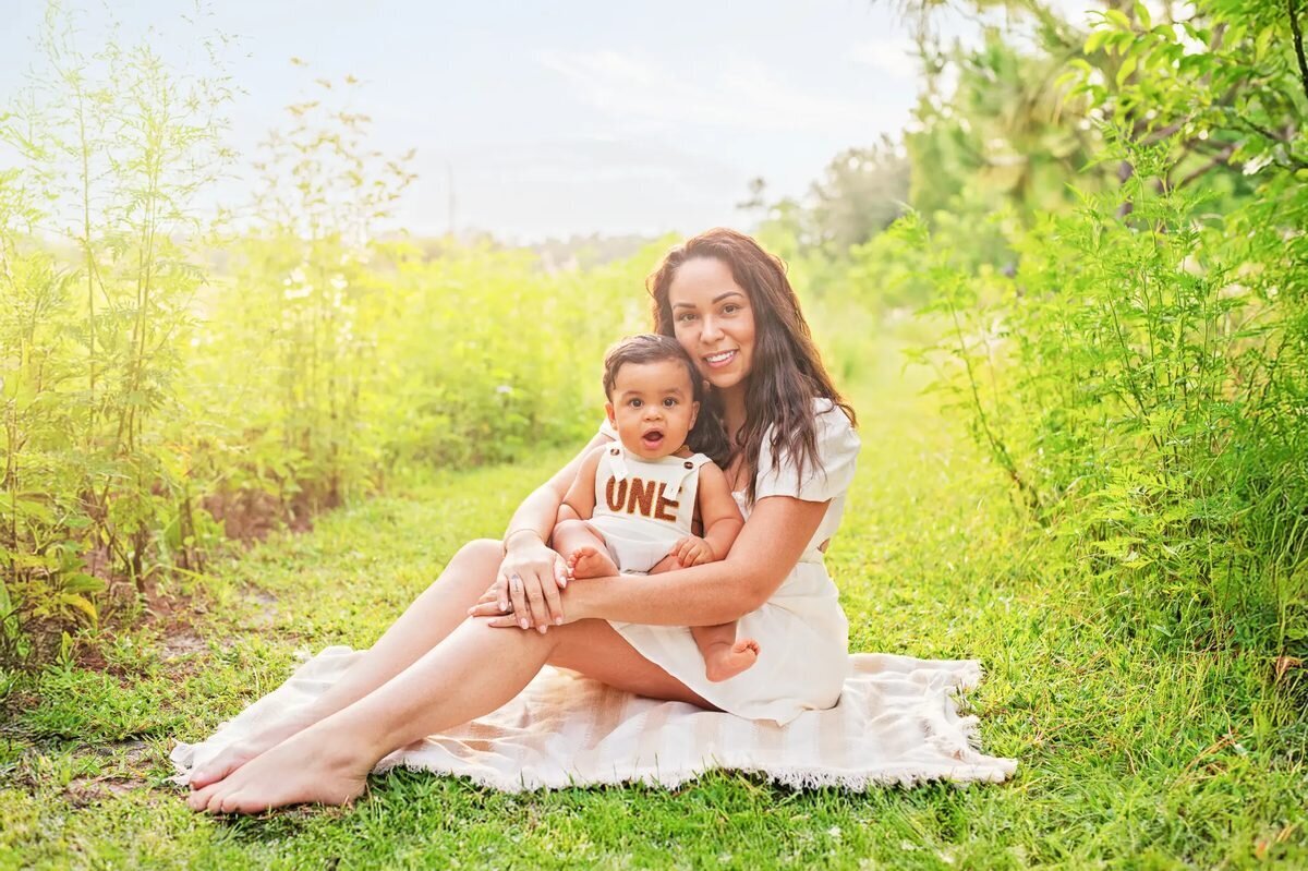 Mother sitting on a blanket in a lush green meadow at Fort De Soto Park, Tampa, holding her baby wearing an "ONE" romper — Tampa family photographer session by Dreamgraphy