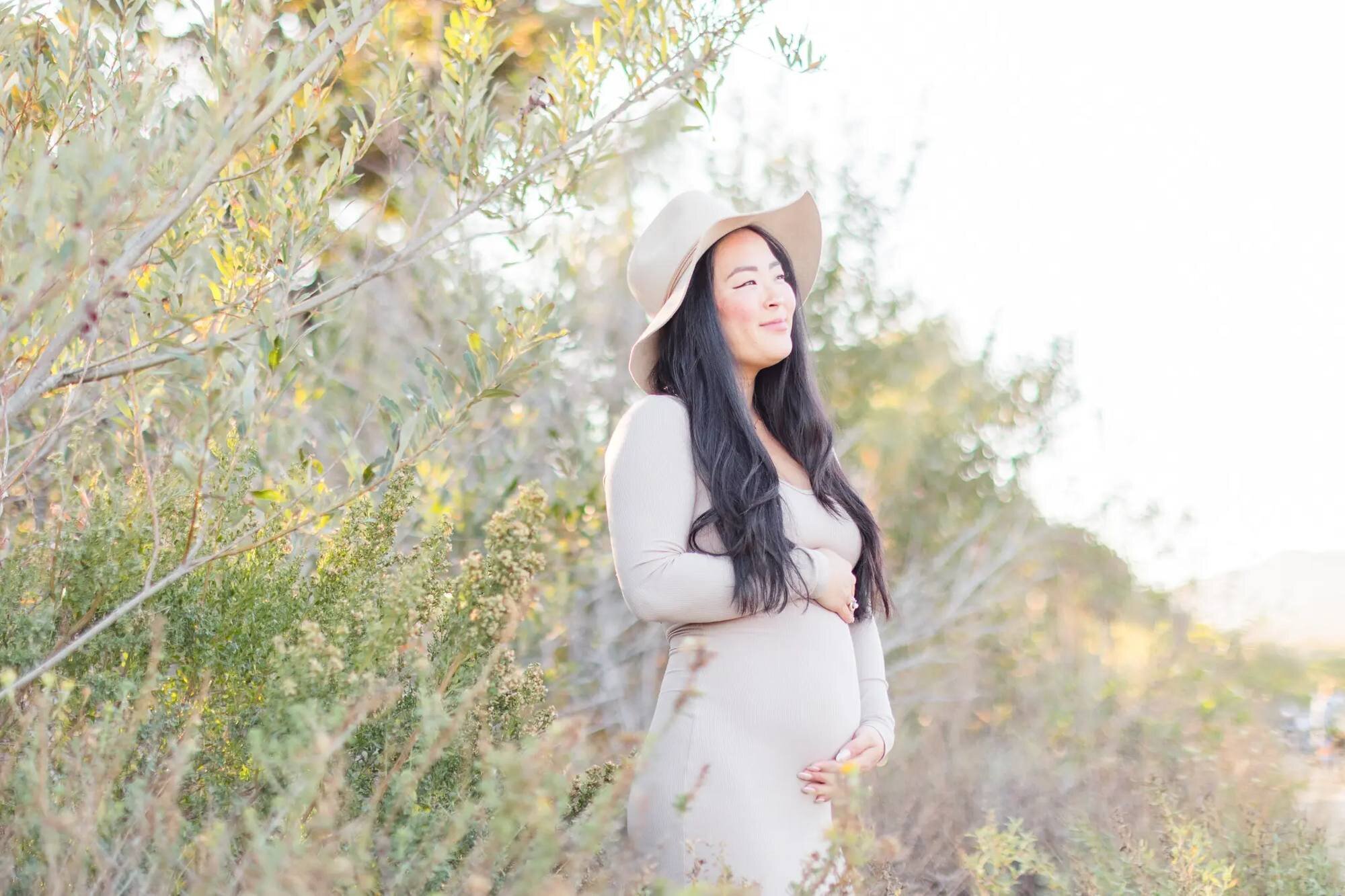 Los Angeles Maternity Photographer — Maternity Session at Malibu Creek State Park Los Angeles maternity photographer capturing a pregnant woman in a beige dress and wide-brim hat cradling her baby bump while standing among golden wildflowers and olive trees at Malibu Creek State Park