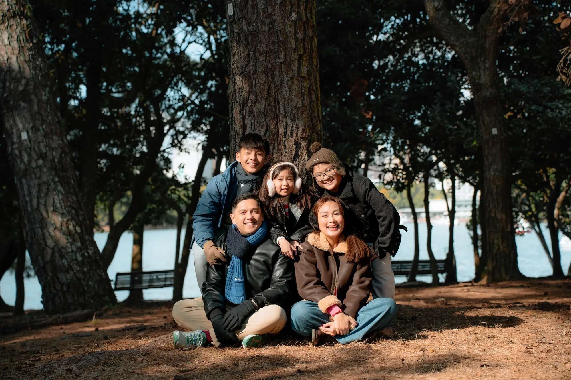 fukuoka-photographer-location-ohori-park A smiling family of five in winter coats gathers under tall pine trees beside the water at Ohori Park in Fukuoka.
