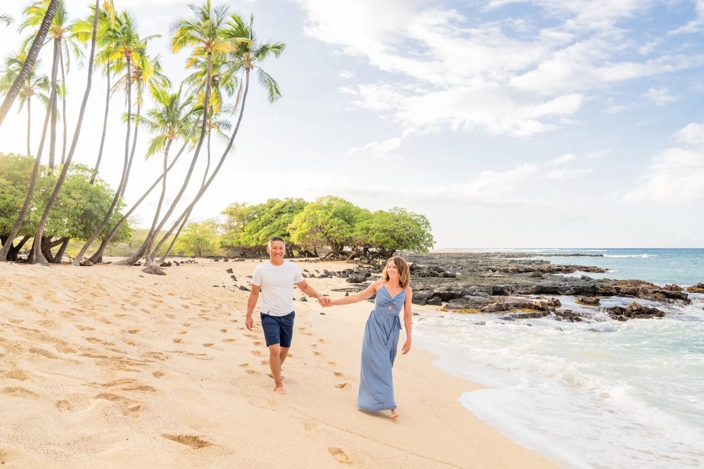 Couple Photos Big Island — Kohala Coast Beach Session A smiling couple walking hand in hand along a golden sand beach on the Big Island with palm trees and turquoise water behind them