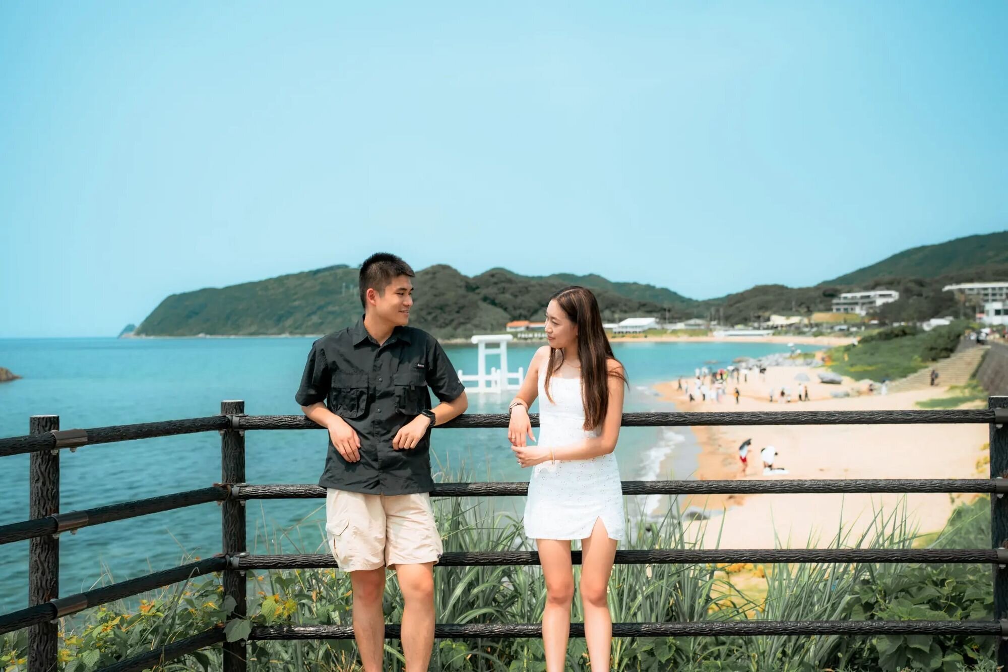 fukuoka-photographer-location-futamigaura-beach A couple leans on a railing above Futamigaura Beach in Itoshima, with a white torii gate standing in the turquoise sea behind them.