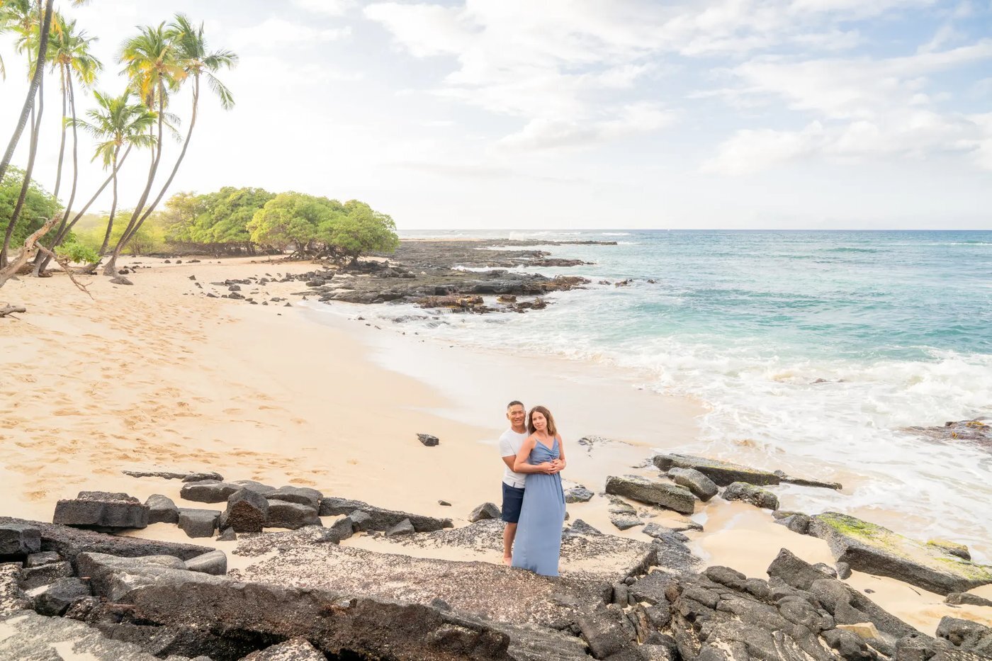 Couple Photoshoot Big Island Hawaii — Lava Rock Beach Portrait A couple embracing on lava rocks on the Big Island of Hawaii with a wide white sand beach, palm trees, and turquoise ocean stretching behind them