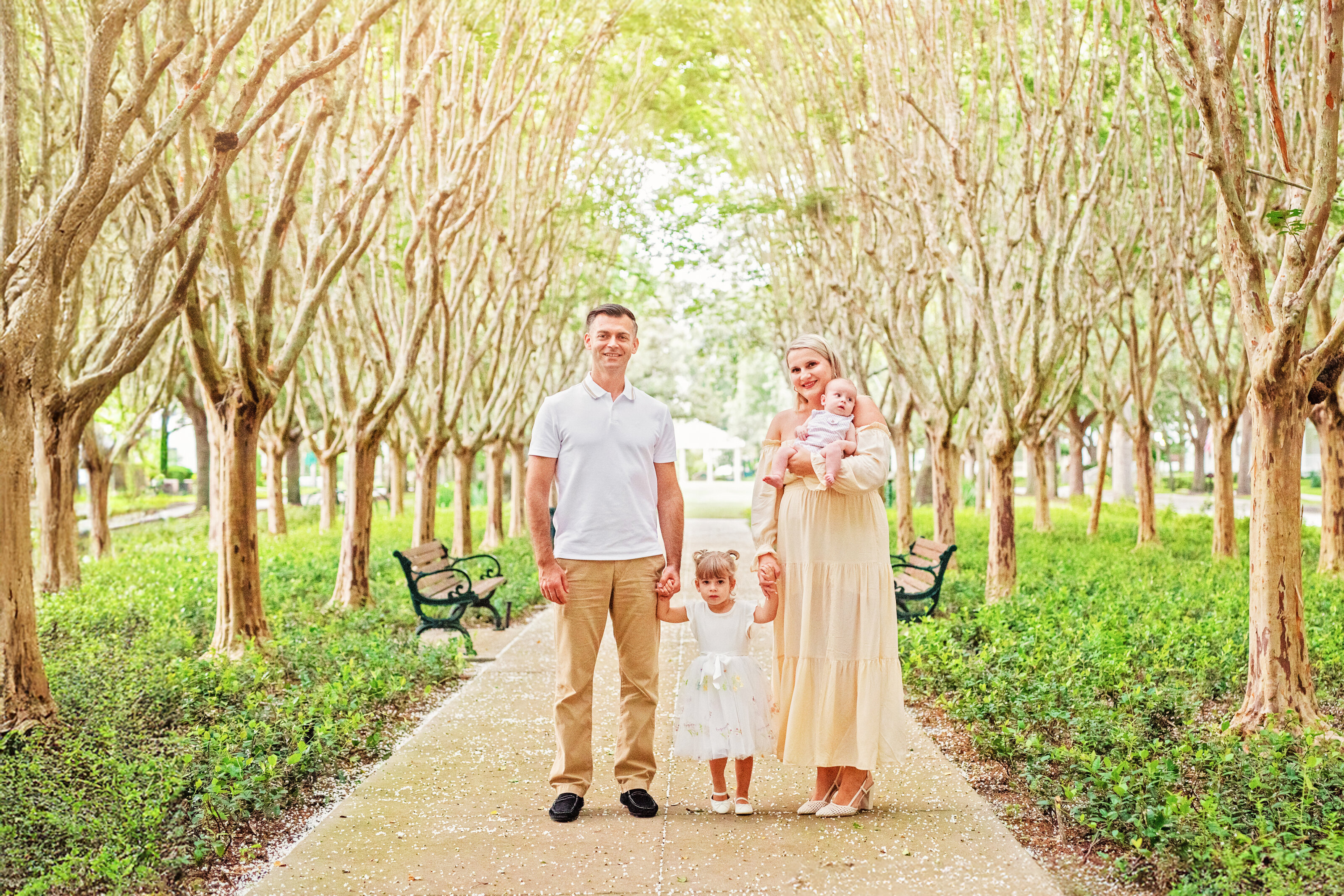 orlando-photographer-family-session-celebration-tree-lined-path-mobile A family of four posing on a tree-lined pathway in Celebration, Florida, with a mother holding a newborn, a toddler daughter between the parents, and lush green gardens and park benches in the background