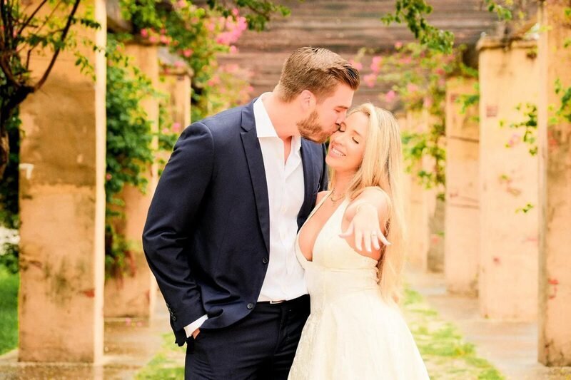 miami-engagement-photographer-vizcaya-mobile A man in a navy suit kissing the cheek of a smiling woman as she shows her ring, among stone pillars and pink bougainvillea in Miami