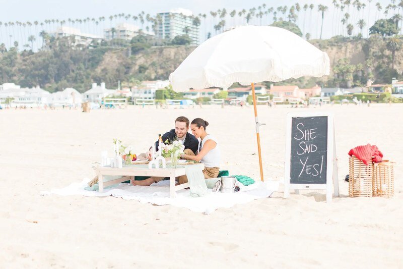 Engagement Proposal Photographer — Surprise Beach Picnic Session by Dreamgraphy A couple celebrating a surprise beach picnic proposal with a She Said Yes sign — engagement proposal photographer by Dreamgraphy