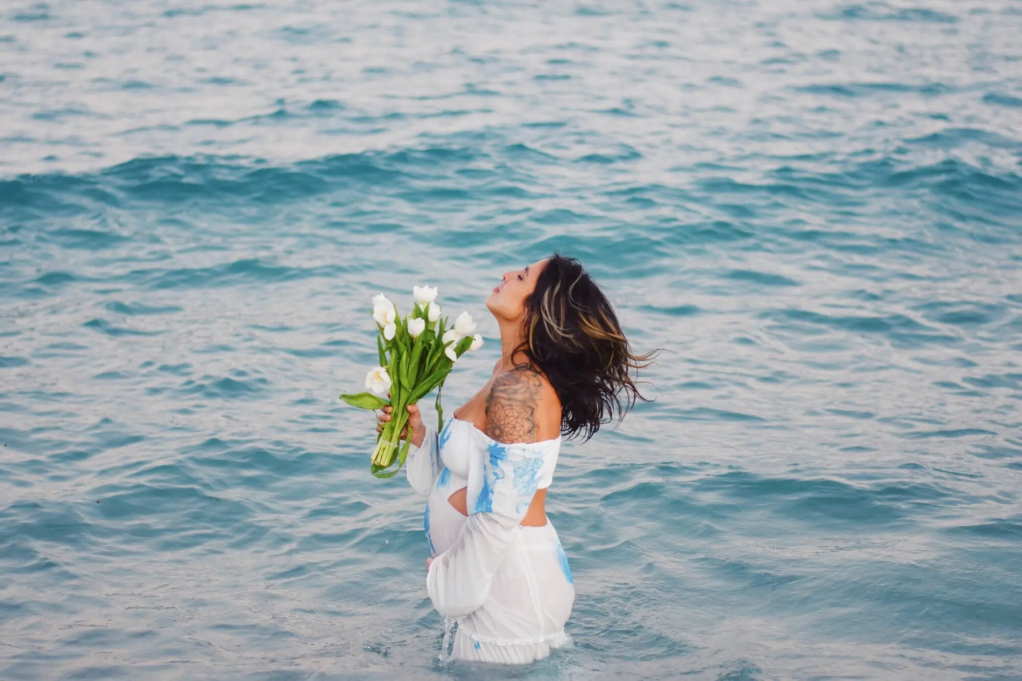 Miami Maternity Photographer | South Beach Ocean Session A pregnant woman standing in the turquoise ocean at South Beach, Miami, holding a bouquet of white tulips with her head tilted back — captured by a Dreamgraphy Miami maternity photographer