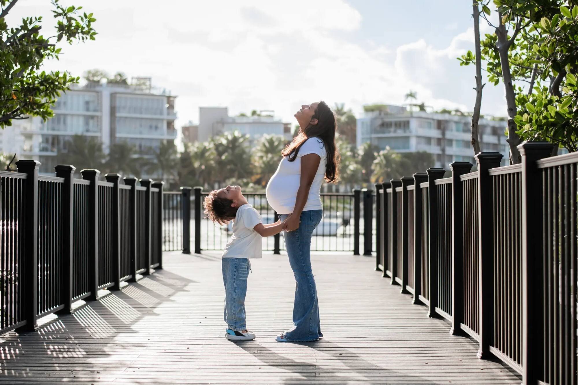 Miami Maternity Photographer | South Pointe Park Boardwalk Session A pregnant woman and her young son standing on the boardwalk at South Pointe Park, Miami, both looking up joyfully — captured by a Dreamgraphy Miami maternity photographer