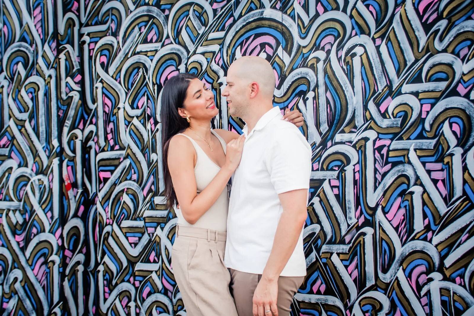 Miami Couple Photographer | Wynwood Graffiti Mural Session A couple gazing at each other and laughing in front of a vibrant graffiti mural in Wynwood, Miami — captured by a Dreamgraphy Miami couple photographer