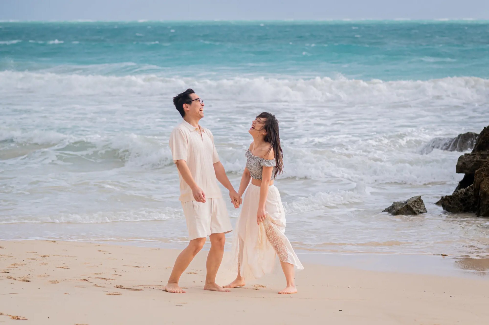 Kauai Photographer — Couple Walking and Laughing on the Beach | Dreamgraphy A couple holding hands and laughing together while walking along a sandy Kauai beach with turquoise ocean waves crashing behind them, captured during a professional couples photo session