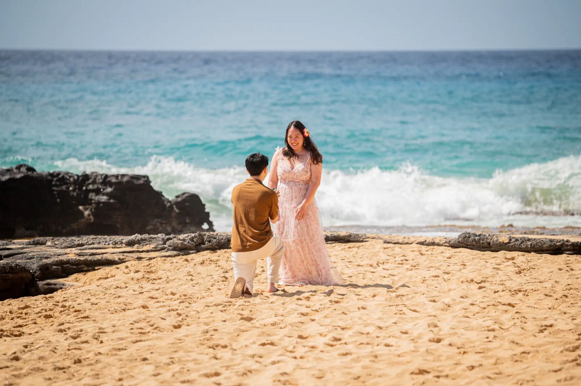 Kauai Photographer — Surprise Beach Proposal Session | Dreamgraphy A man proposing on one knee on a sandy Kauai beach as his partner reacts with joy, with turquoise ocean waves and volcanic rocks in the background, captured by a professional Kauai proposal photographer