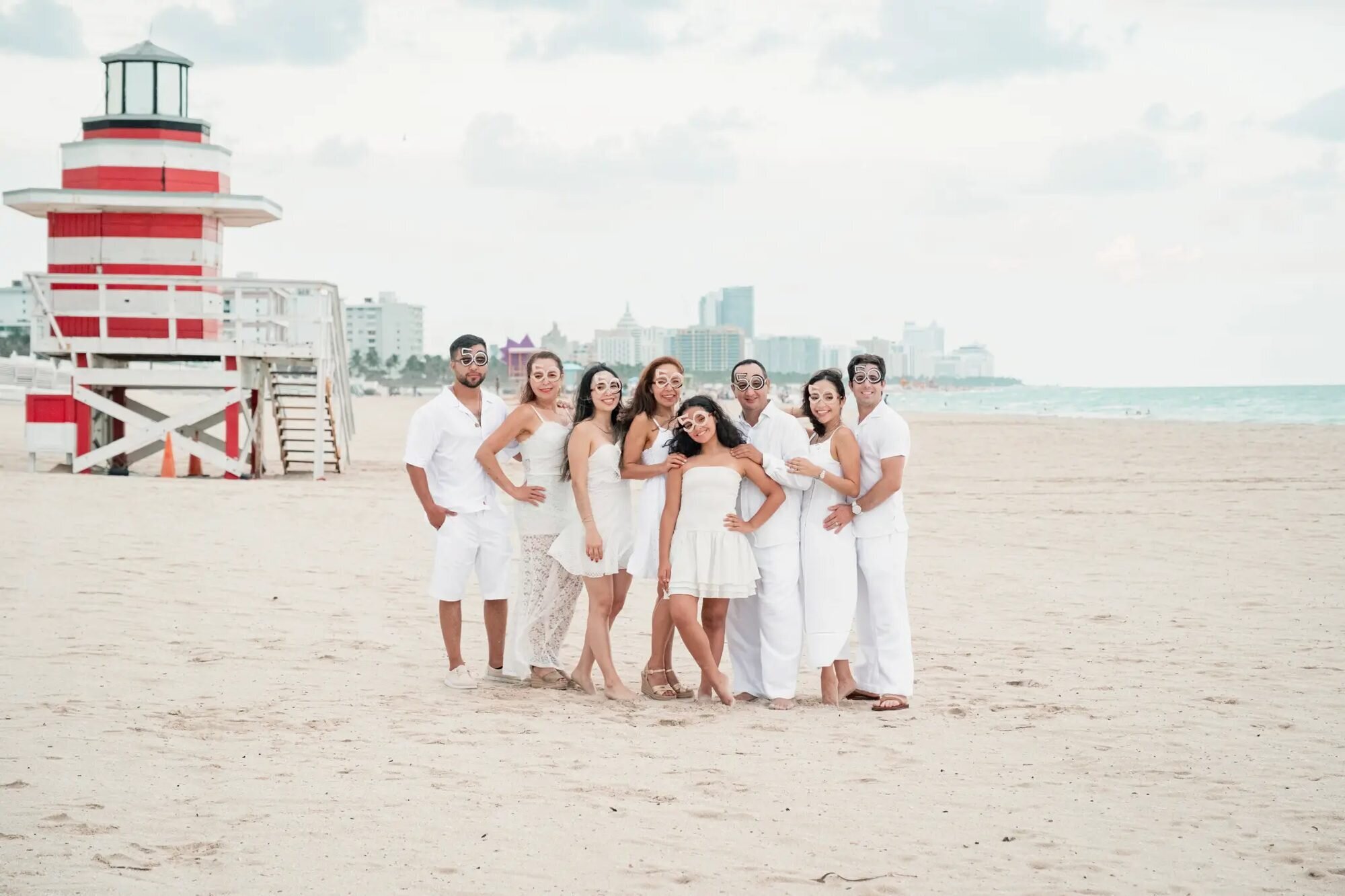 Miami Family Photographer | Group Family Session at South Beach A large family of eight dressed in white laughing together on the sand at South Beach, Miami, with the iconic red-and-white lifeguard tower in the background — captured by a Dreamgraphy Miami family photographer