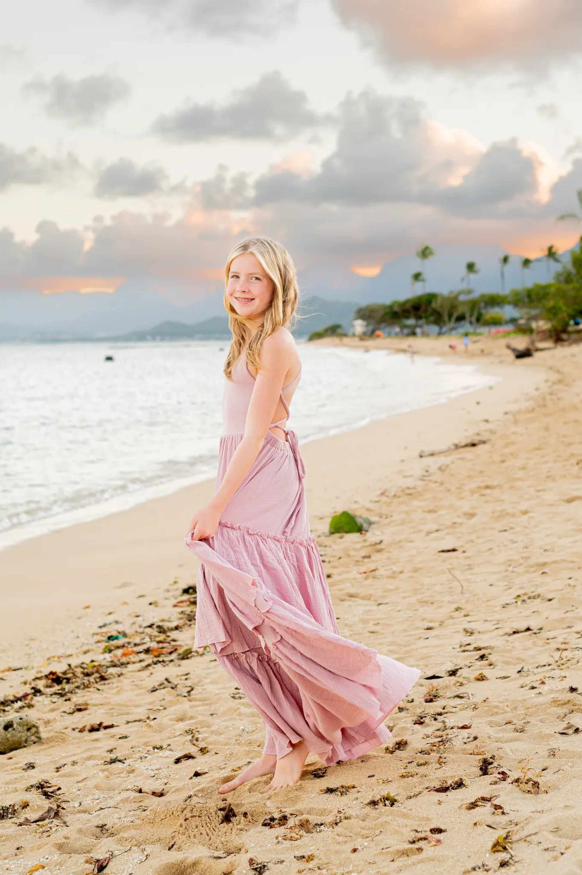 Kauai Photographer — Family Beach Sunset Portrait | Dreamgraphy A young girl in a flowing pink dress twirling on a sandy Kauai beach at golden hour, with soft waves and a pastel sunset sky in the background, captured during a professional family photo session