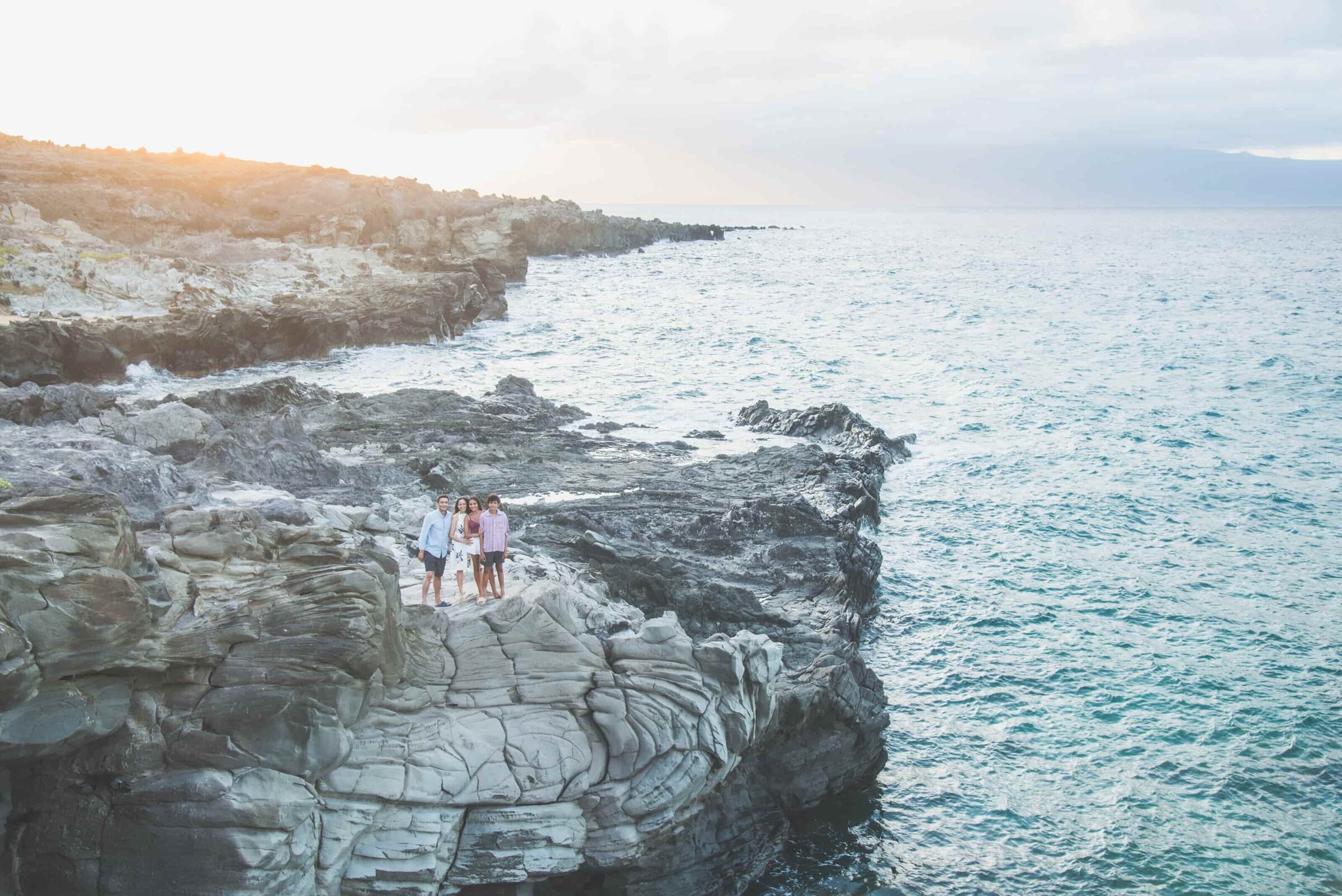 A family of four standing together on wide grey lava-rock cliffs above the ocean, with the sun low over the headland, on Maui