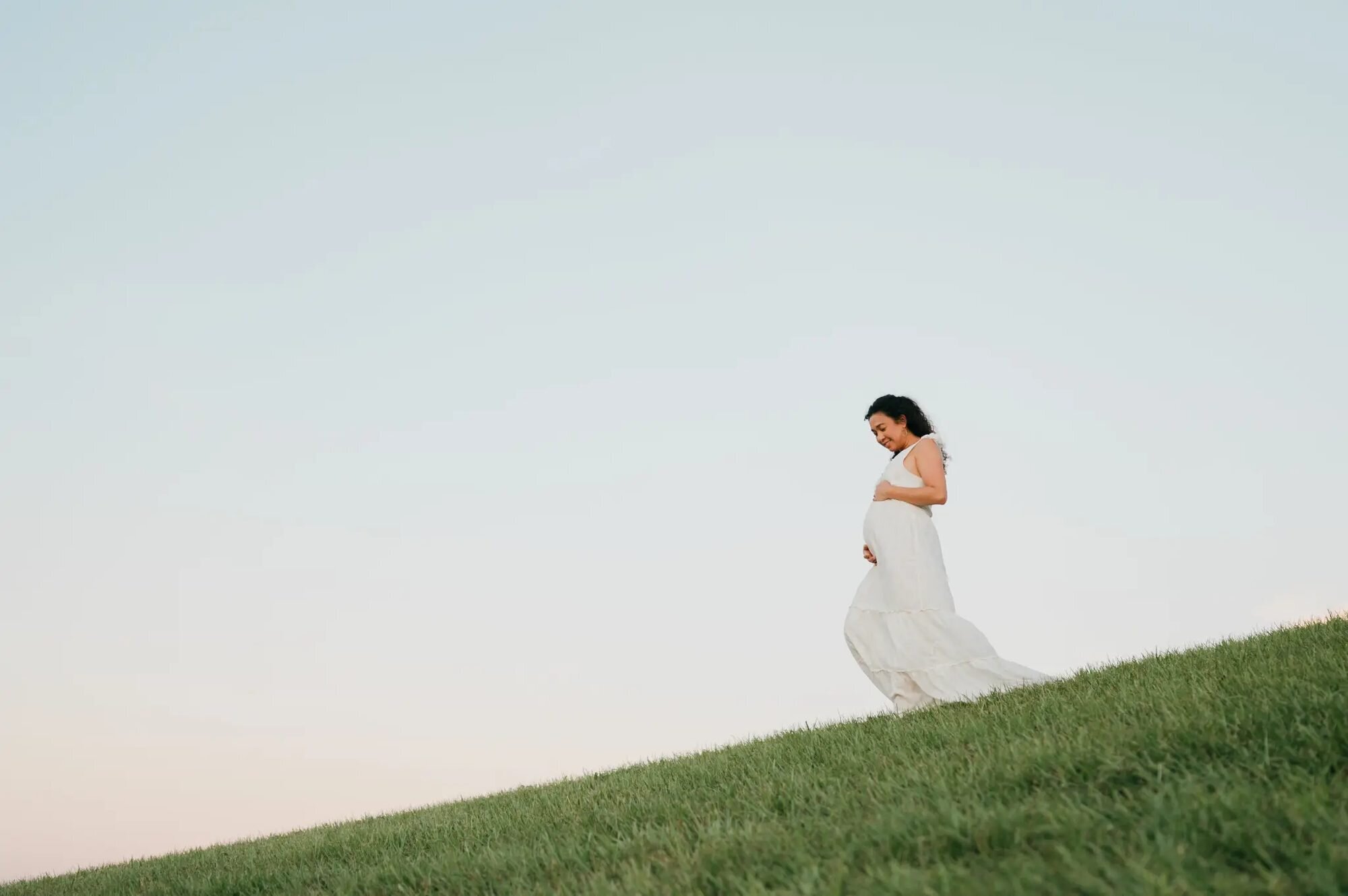 Tampa Maternity Photographer | Outdoor Golden Hour Session A pregnant woman in a flowing white dress cradling her baby bump while standing on a grassy hillside at golden hour in Tampa, Florida — captured by a Dreamgraphy Tampa maternity photographer