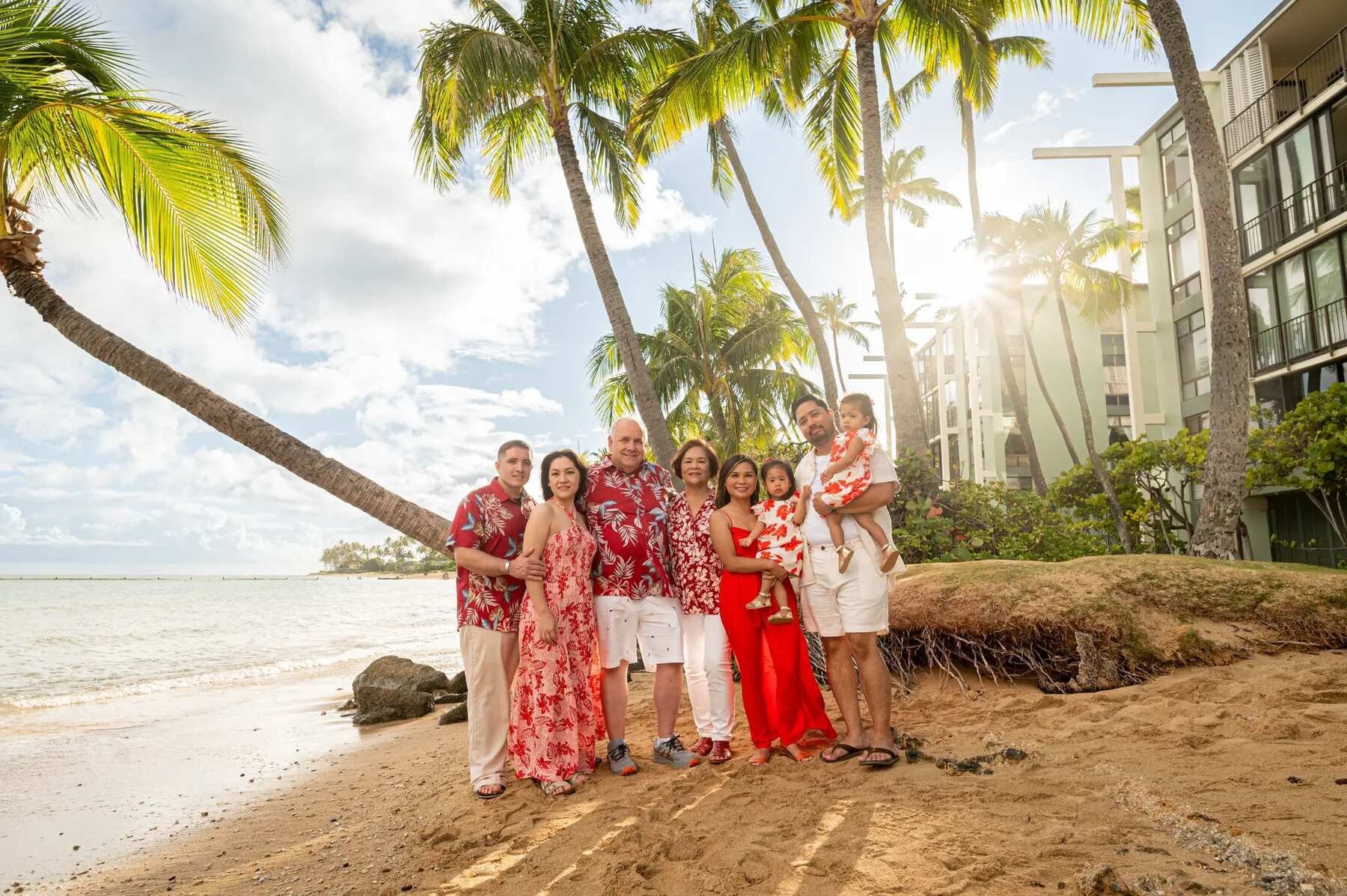 An extended family of eight posing together at Waialae Beach on Oahu with tall palm trees, golden sunlight, and the ocean behind them