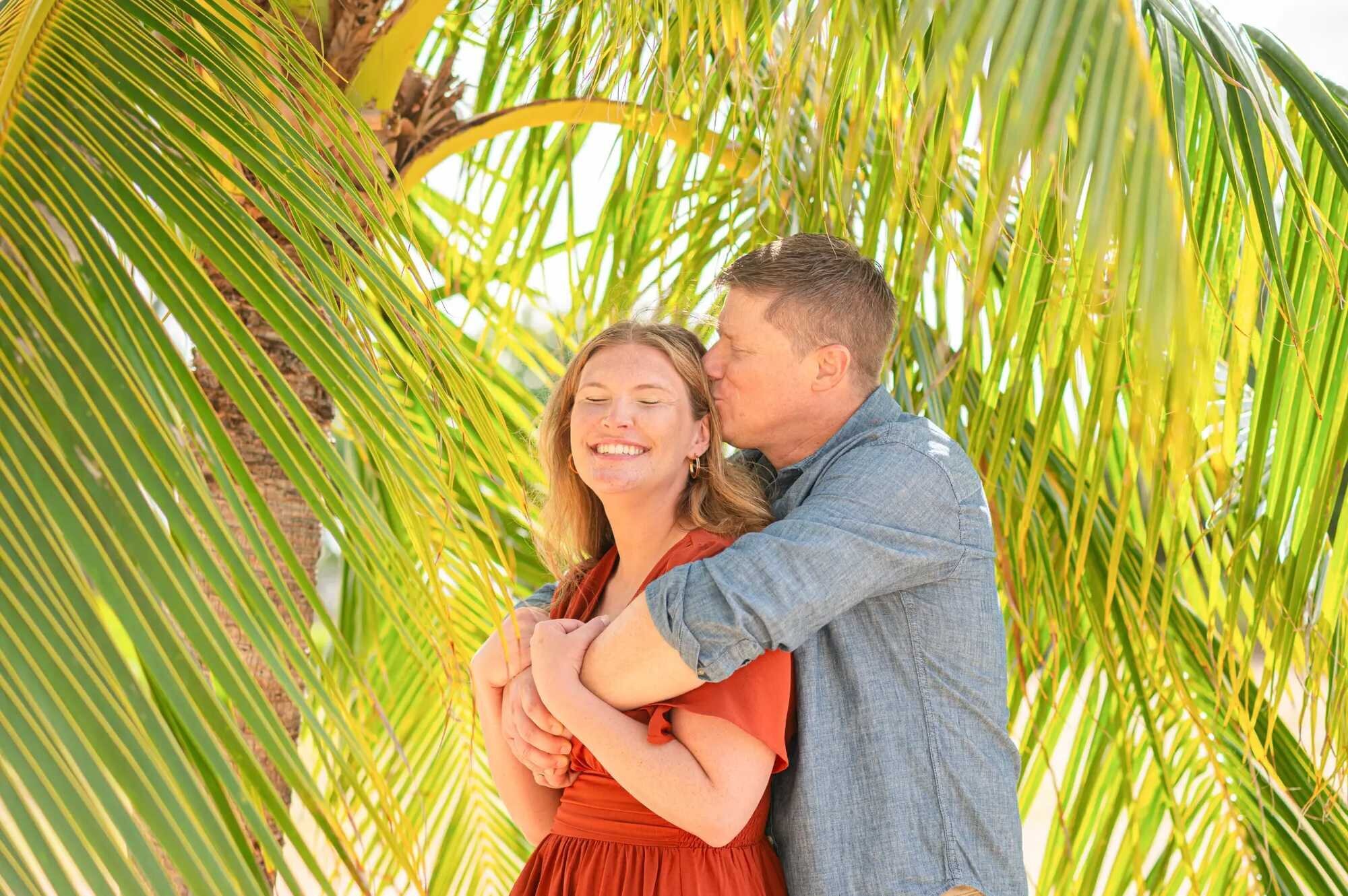 Man kissing a smiling woman on the cheek beneath tropical palm fronds