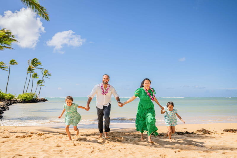 A family of four laughing and running hand in hand along a sandy Hawaii beach with palm trees and turquoise water in the background during a vacation photography session