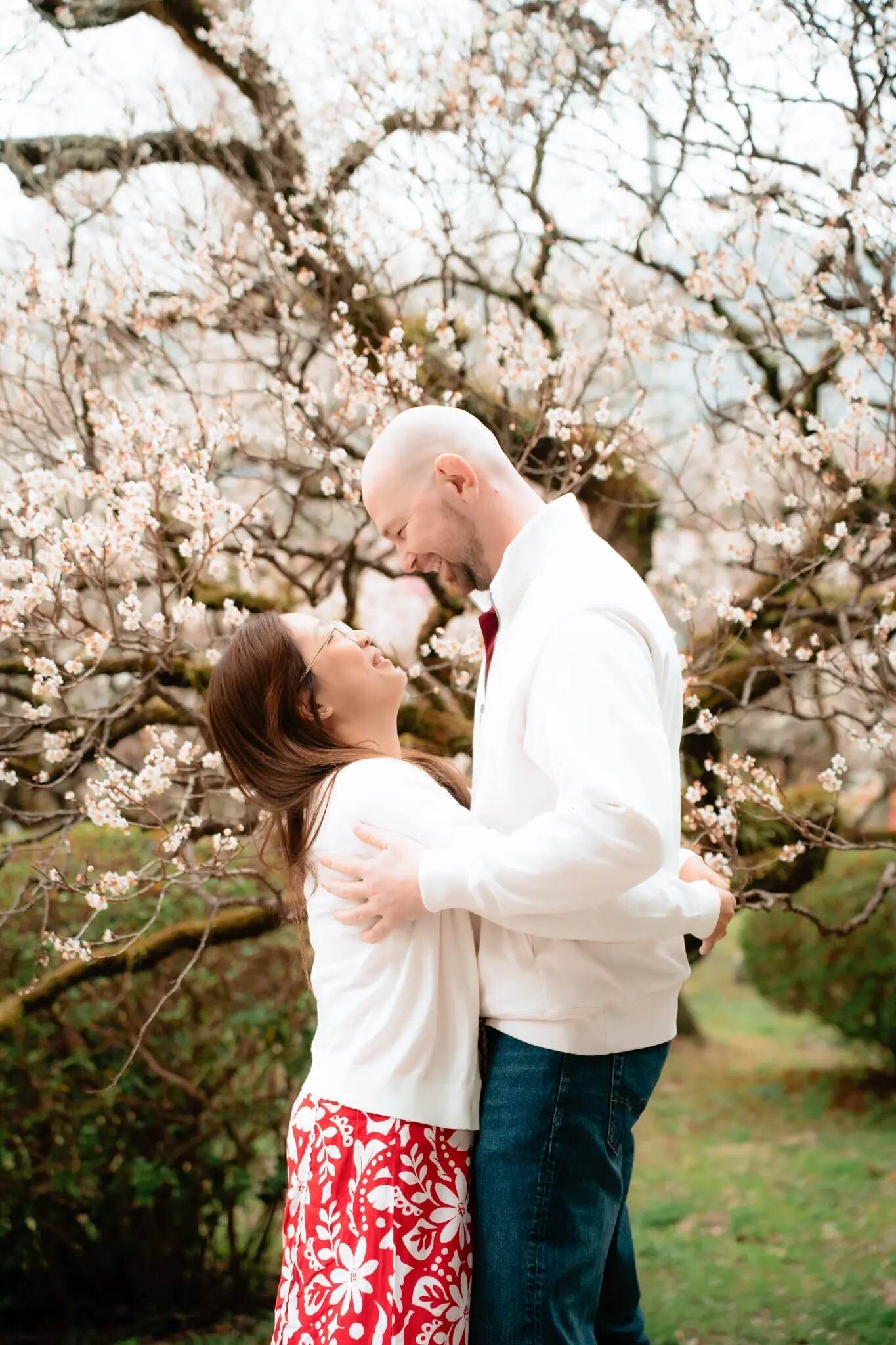 fukuoka-photographer-location-uminonakamichi-seaside-park A laughing couple in white tops embraces beneath a tree covered in pale blossoms at Uminonakamichi Seaside Park in Fukuoka.