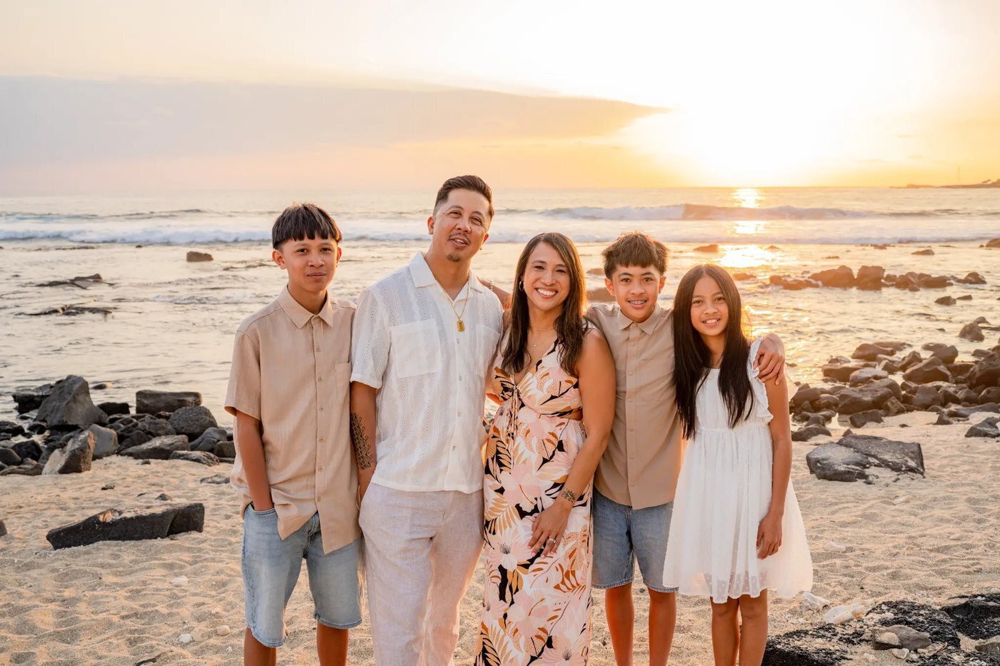 Big Island Family Photographer — Beach Sunset Session Kohala Coast A family of five smiling together on a sandy Big Island beach at golden sunset with lava rocks and ocean waves behind them