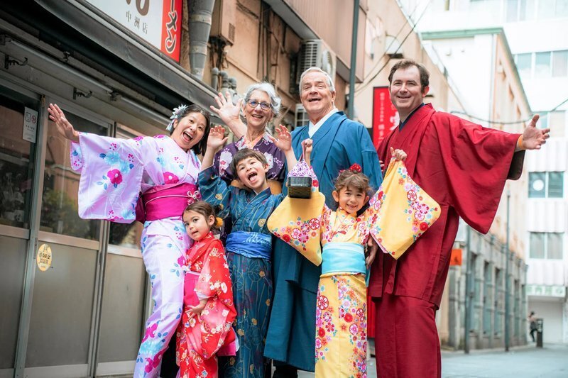 fukuoka-photographer-location-nakasu-mobile Seven adults and children in colorful kimono cheer and wave on a narrow street lined with shop signs in Nakasu, Fukuoka.
