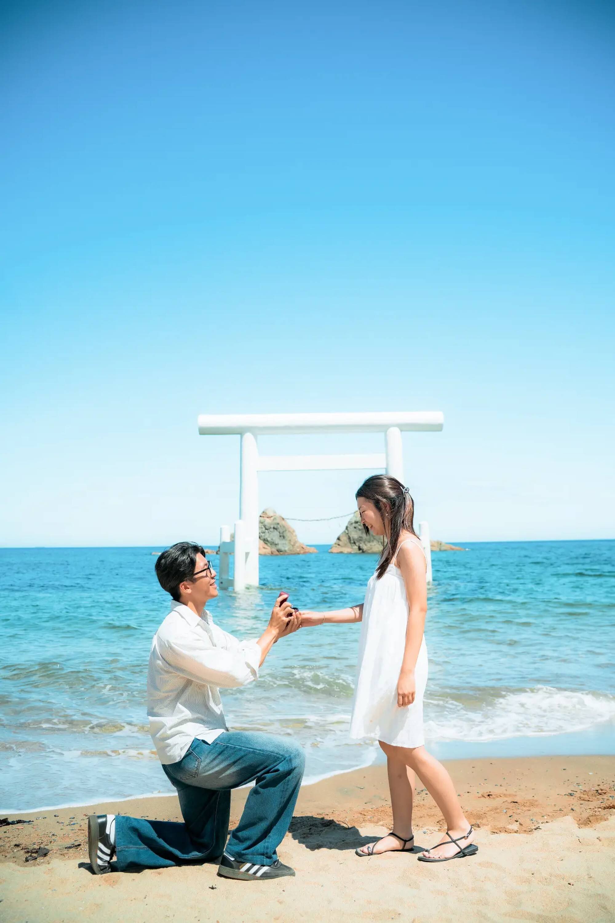 fukuoka-photographer-beach-proposal-session A man on one knee offers a ring to a smiling woman in a white sundress on a beach in Fukuoka, with a white torii gate standing in the sea