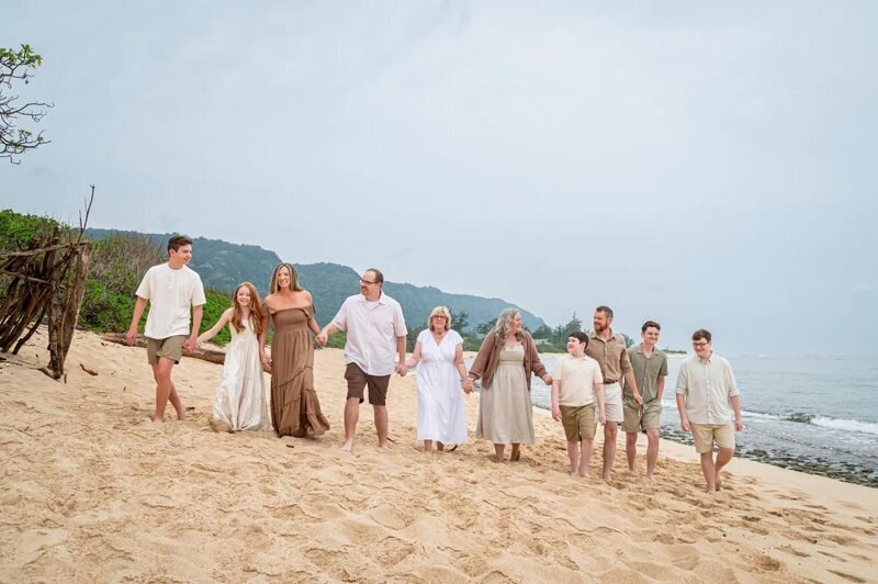 Extended multi-generational family of ten walking hand-in-hand along a sandy Kauai beach with lush green mountains in the background — Kauai family photographer session by Dreamgraphy