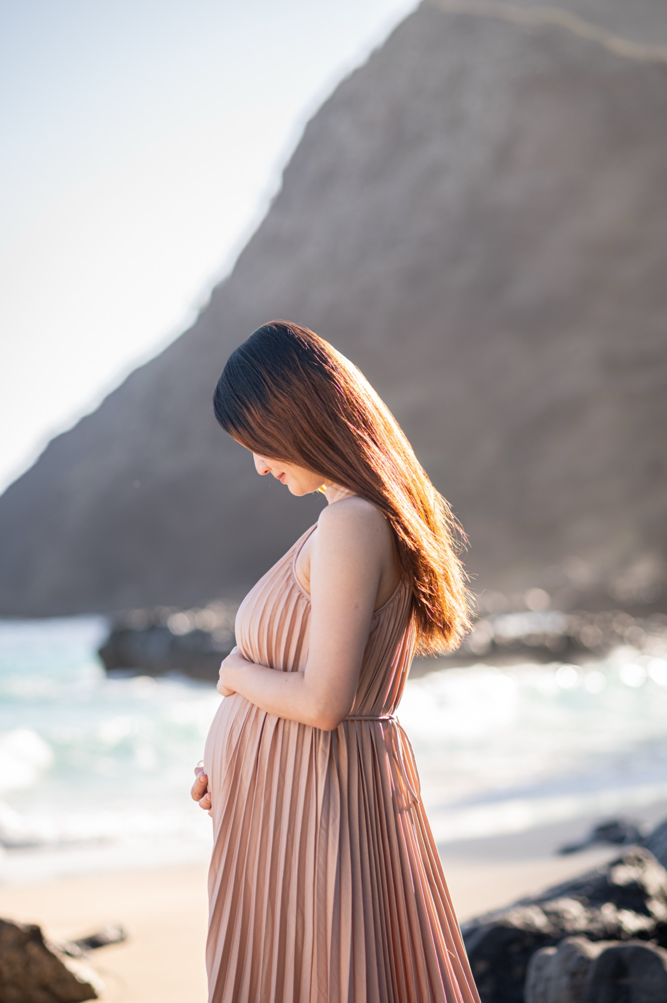 A pregnant woman in profile on the beach, looking down at her bump with her hands resting on it, the shore break soft behind her