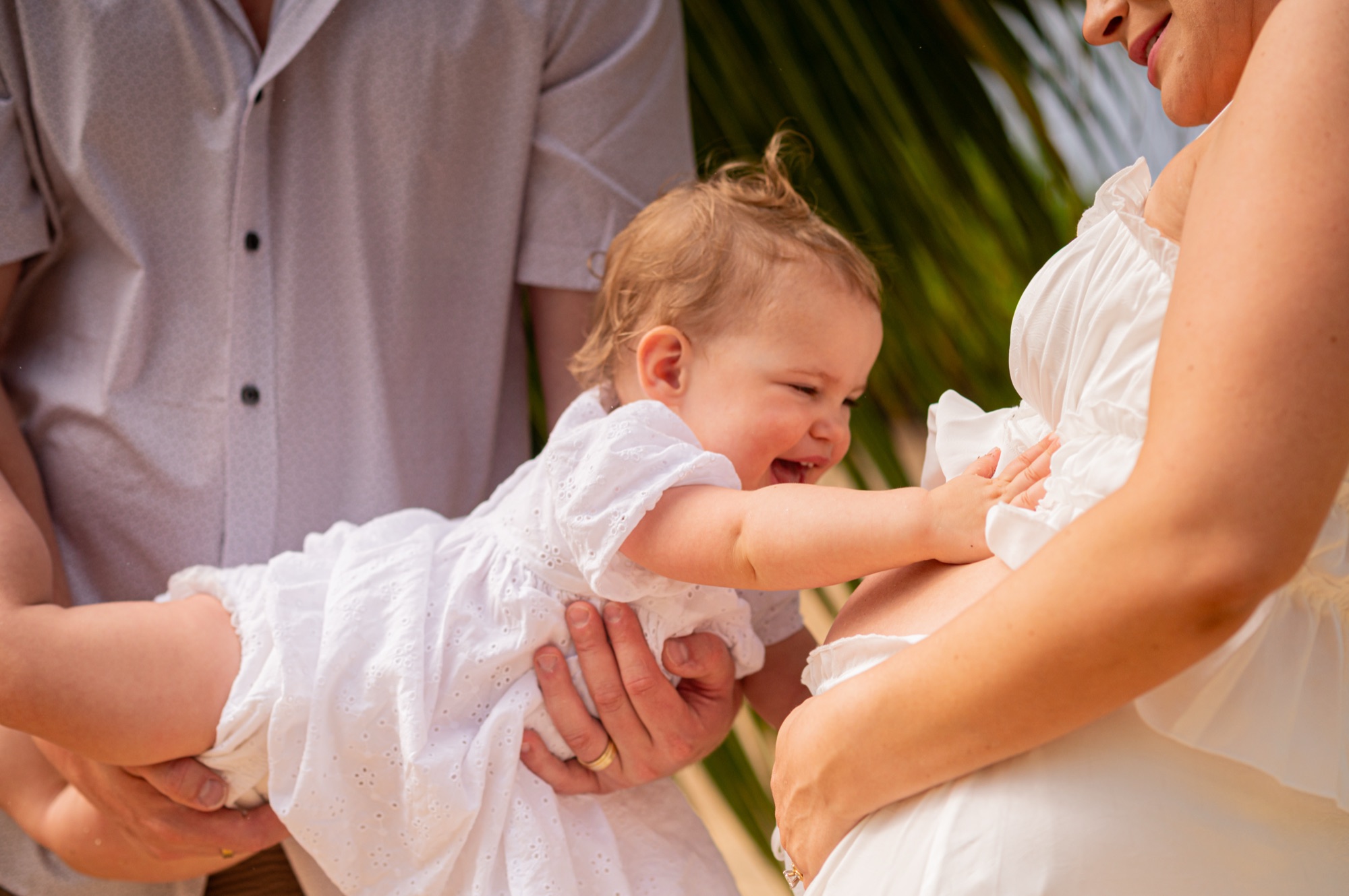 A laughing baby girl reaching out to touch her mother's pregnant bump while her dad holds her, on a beach