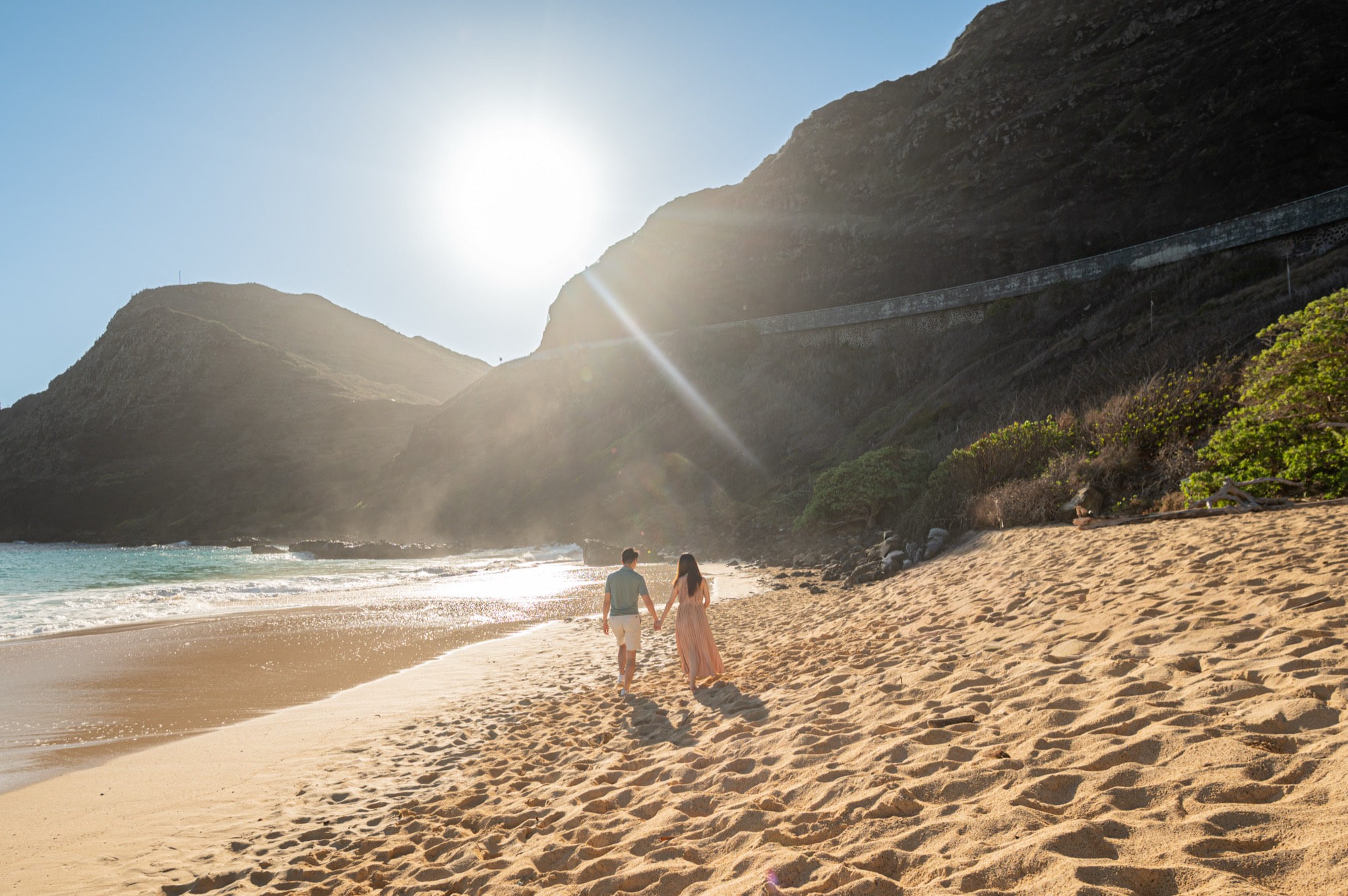 A pregnant woman and her husband walking hand in hand along the sand at the water's edge, with sunlight streaming over the cliffs