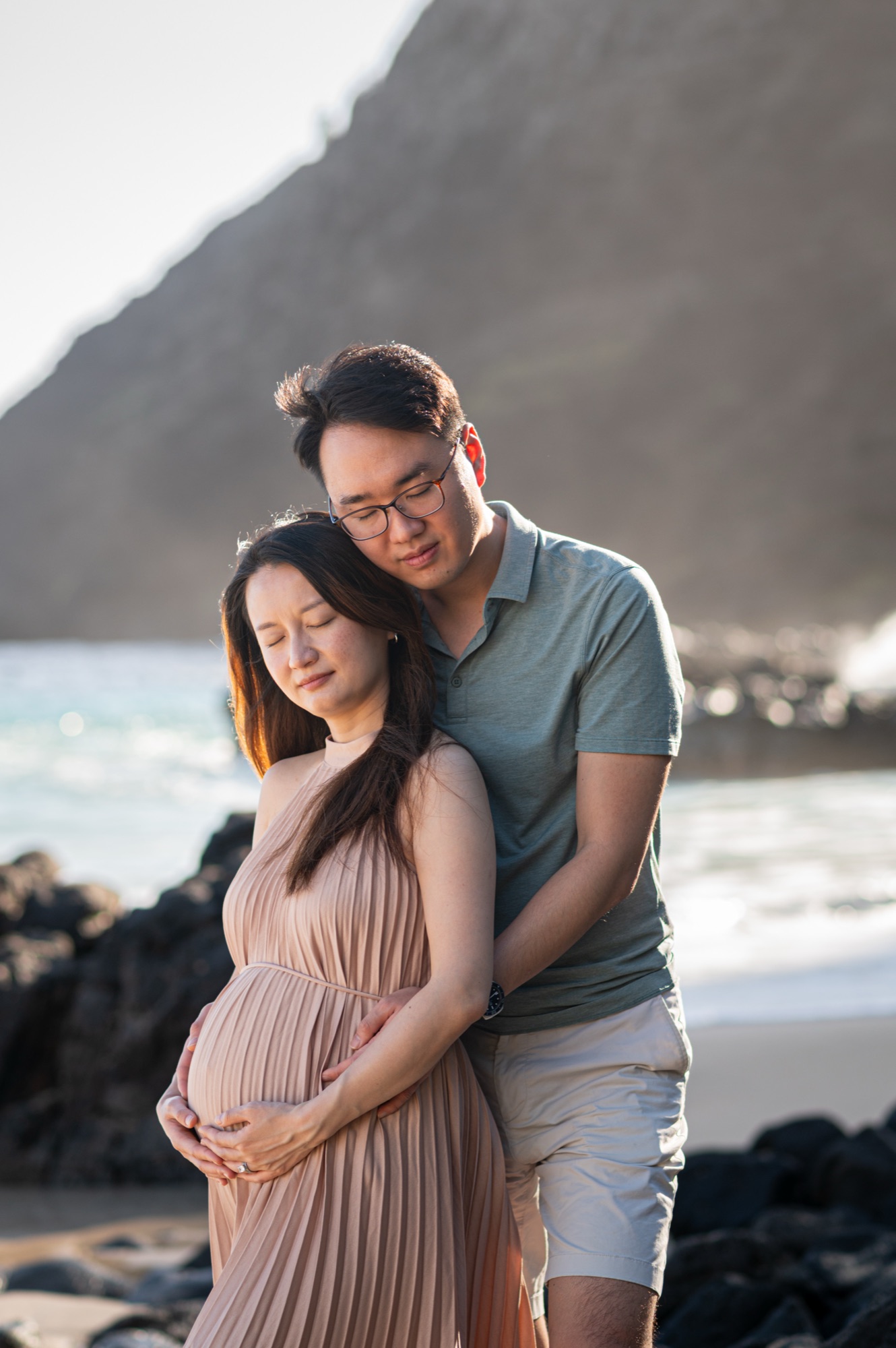 A husband hugging his pregnant wife from behind on a beach, both holding her bump, eyes closed, with a mountain and ocean behind them