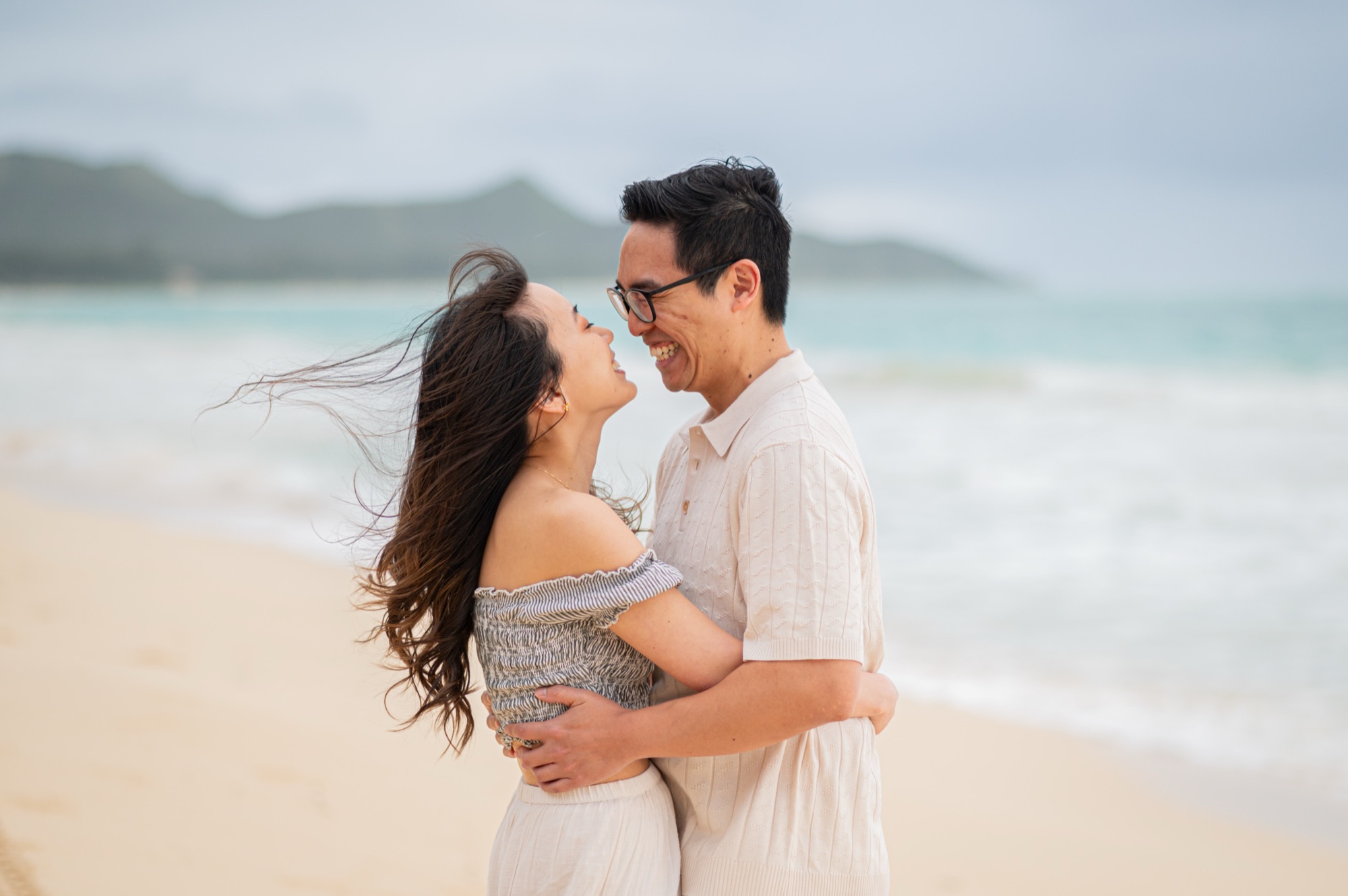 A couple in an almost-kiss on the beach, noses touching and both smiling, her hair blowing in the sea breeze
