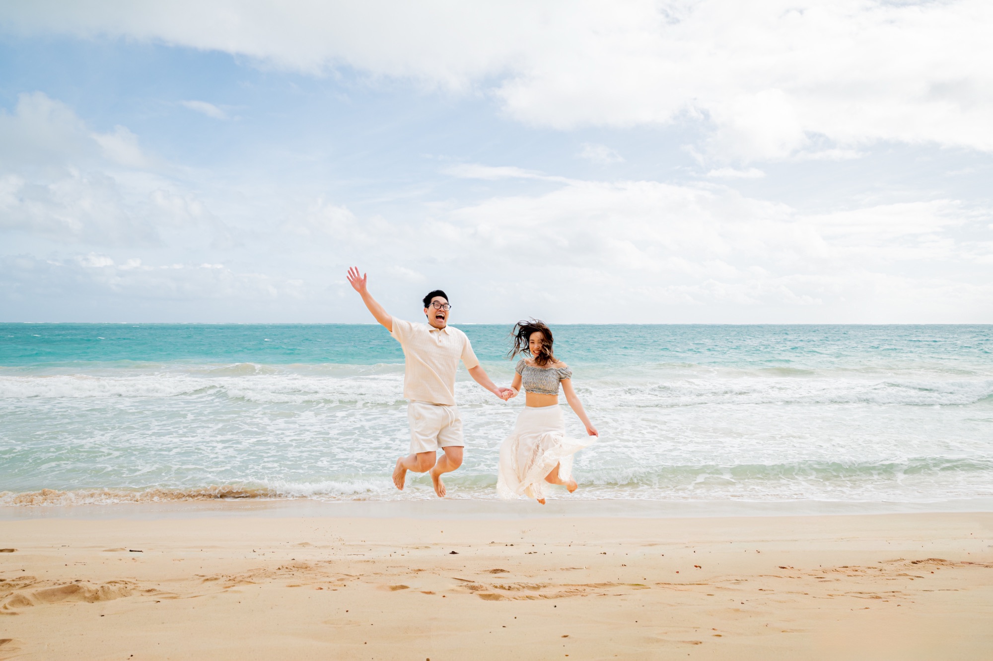 A couple jumping in the air hand in hand on a sandy beach, turquoise waves and a bright cloudy sky behind them