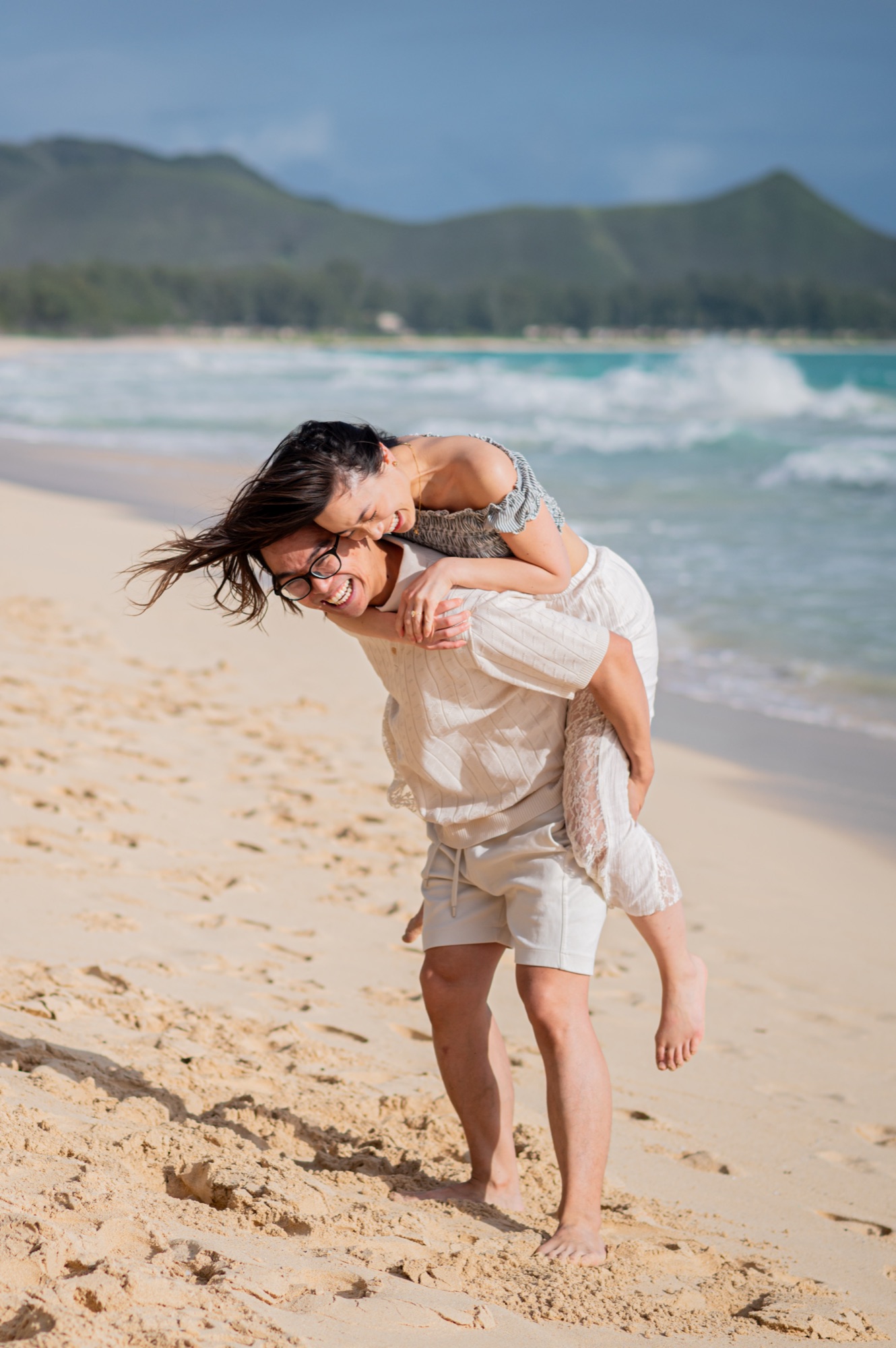 A laughing woman riding piggyback on her partner's back on the beach, the ocean and green hills behind them