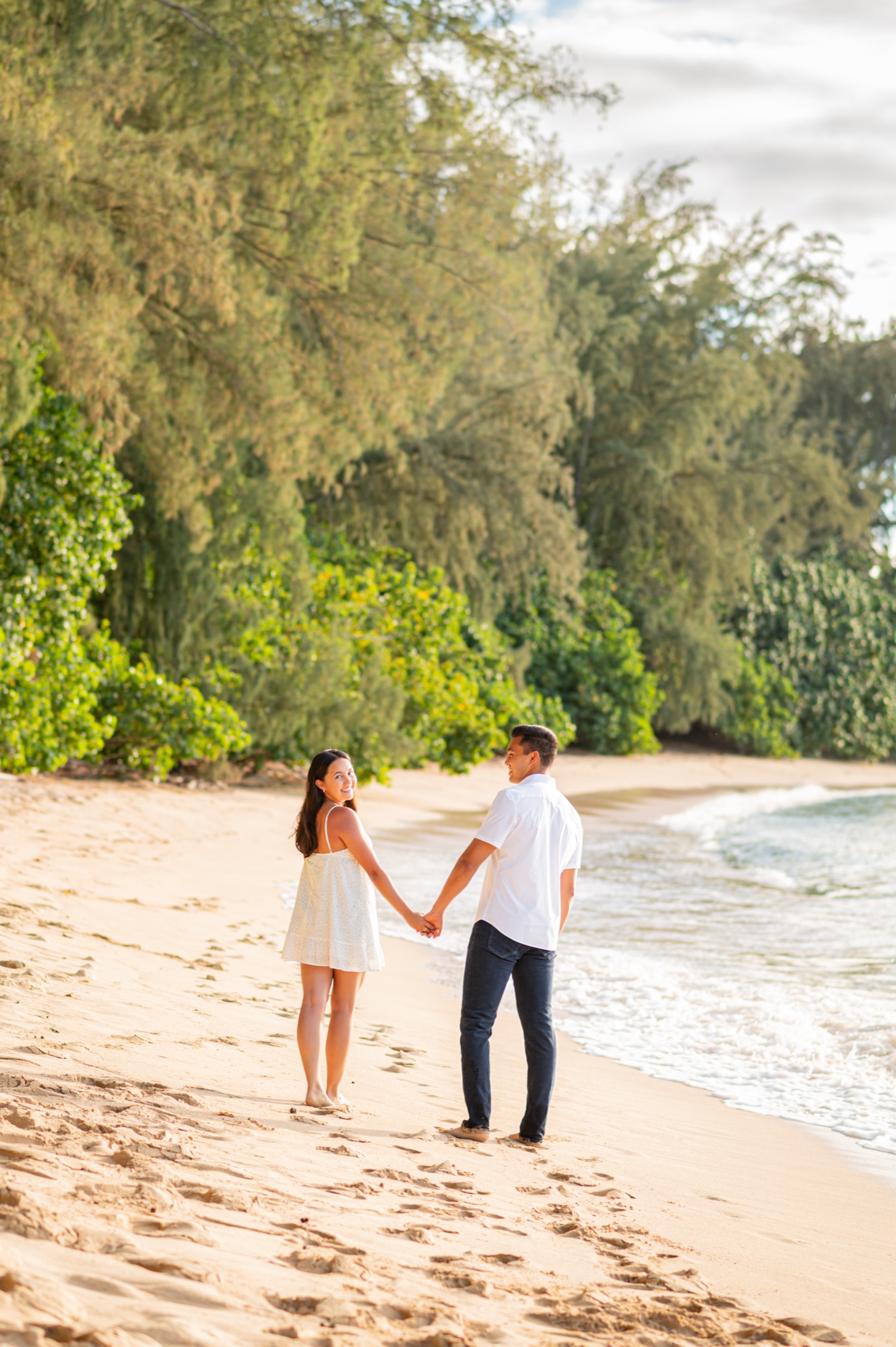 A couple walking hand in hand along the shoreline under tall trees, the woman glancing back over her shoulder