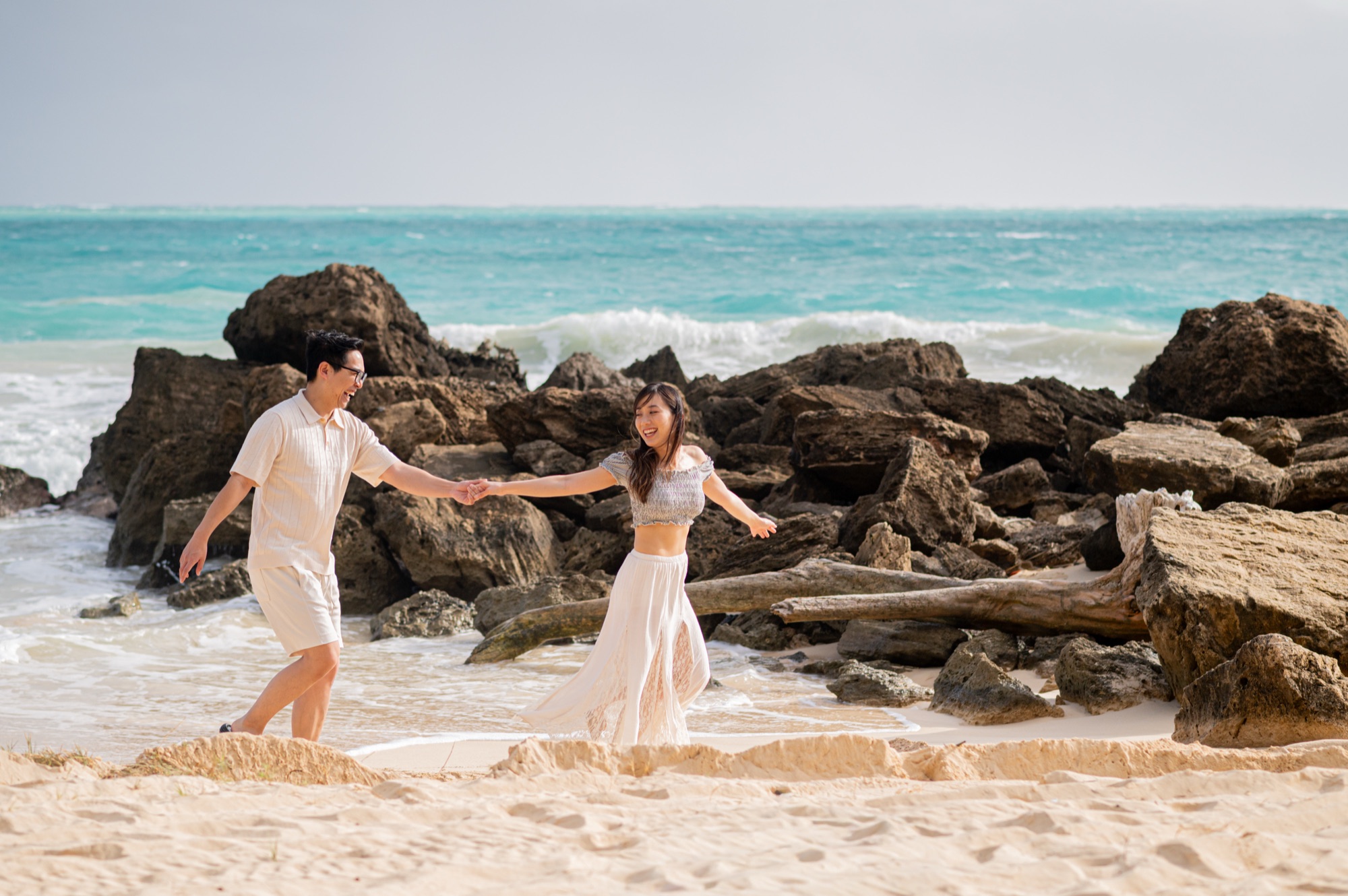 A couple running hand in hand across the sand past dark rocks, turquoise waves breaking behind them