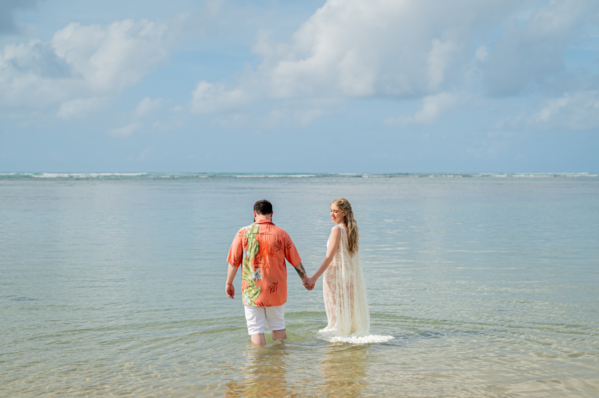A couple wading hand in hand into calm, clear ocean water, the woman in a flowing white dress smiling at her partner